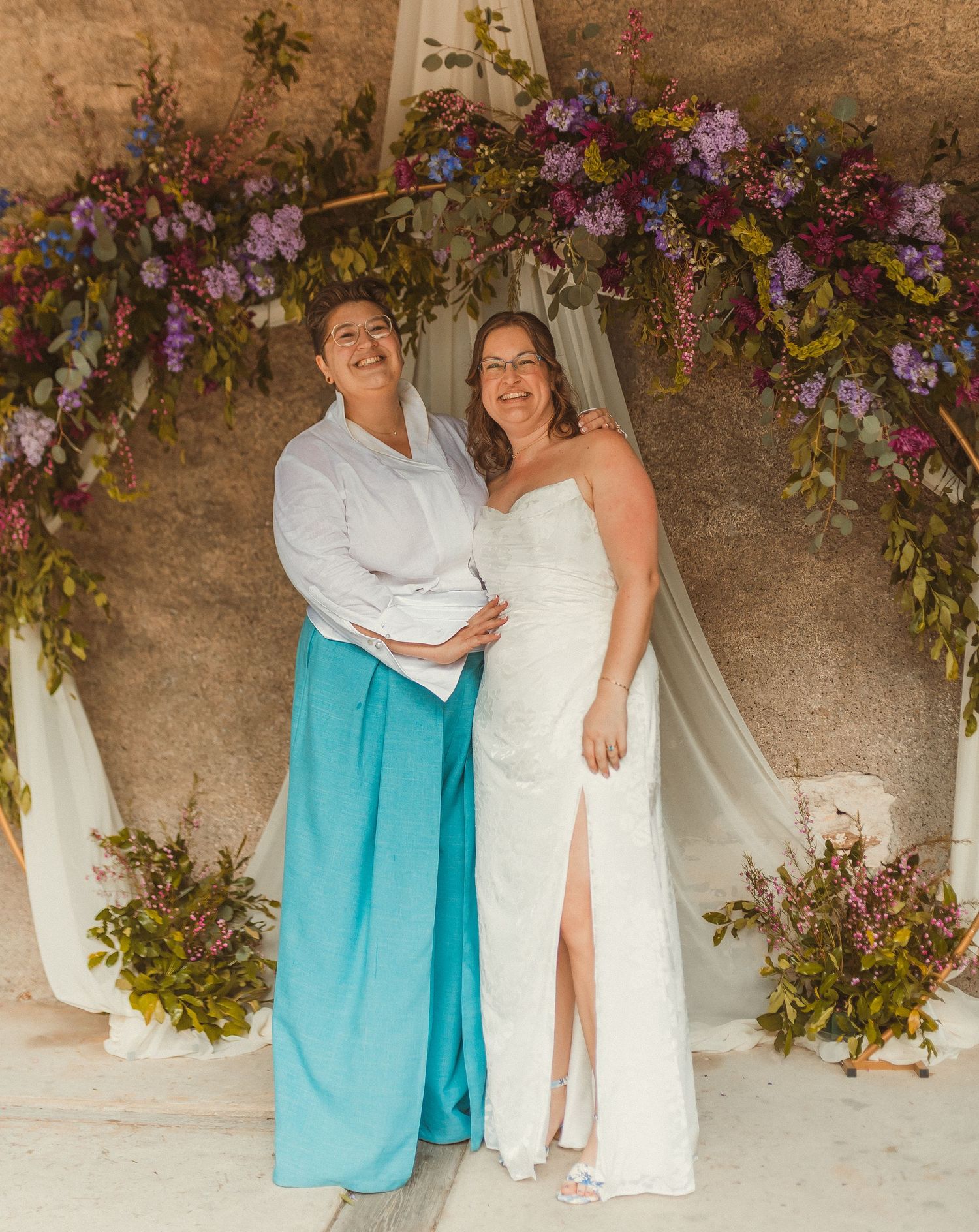 A wedding couple poses under a beautiful floral arch with purple and pink blooms.