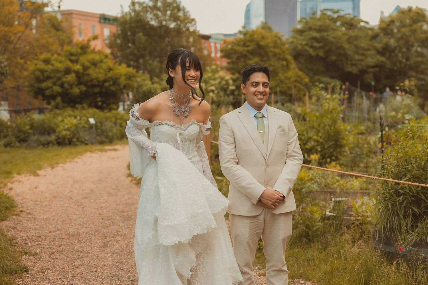 A couple in wedding attire walks together along a dirt path with city buildings visible in the background.