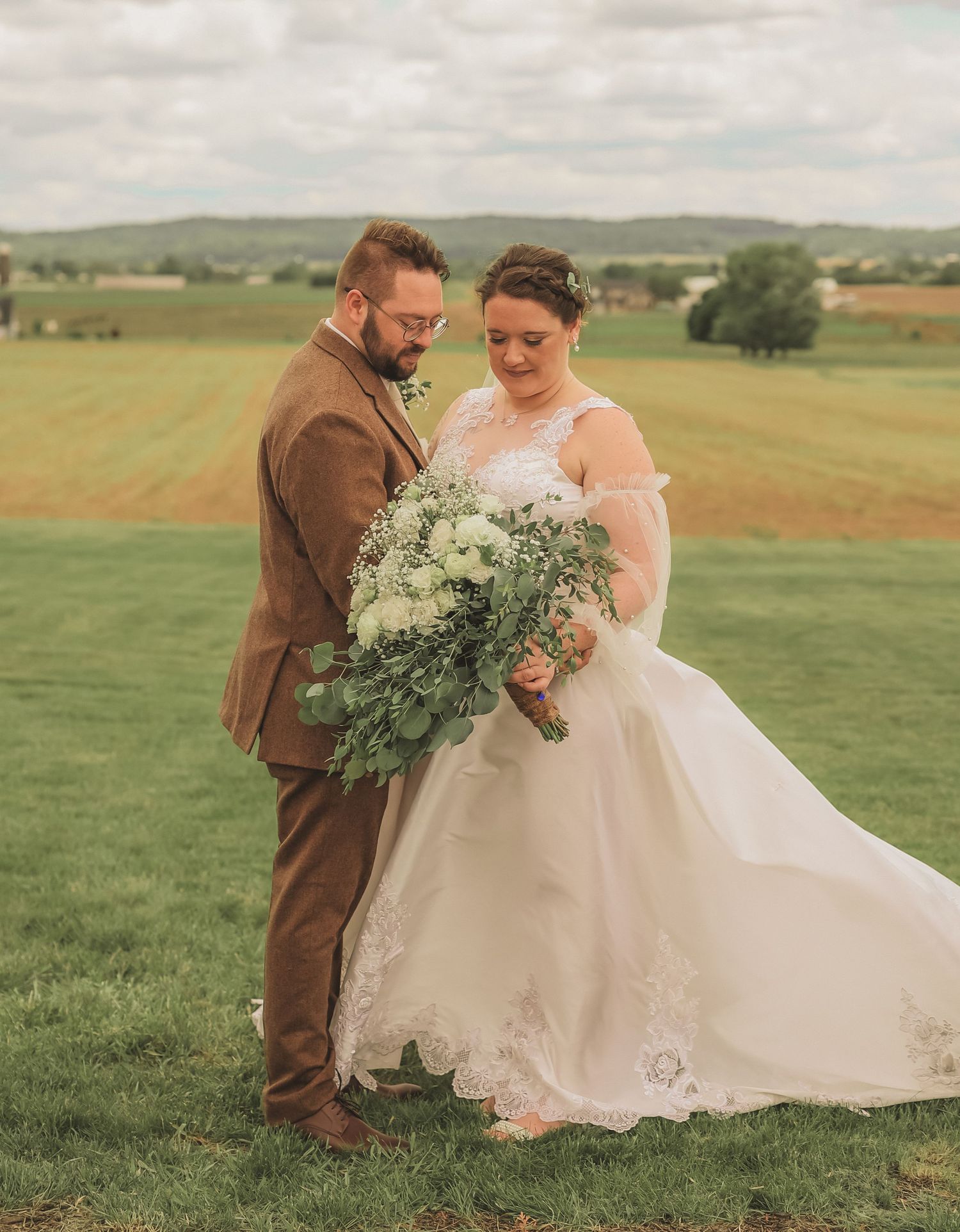 A couple admires a lush green and white wedding bouquet against rolling countryside hills on their special day.