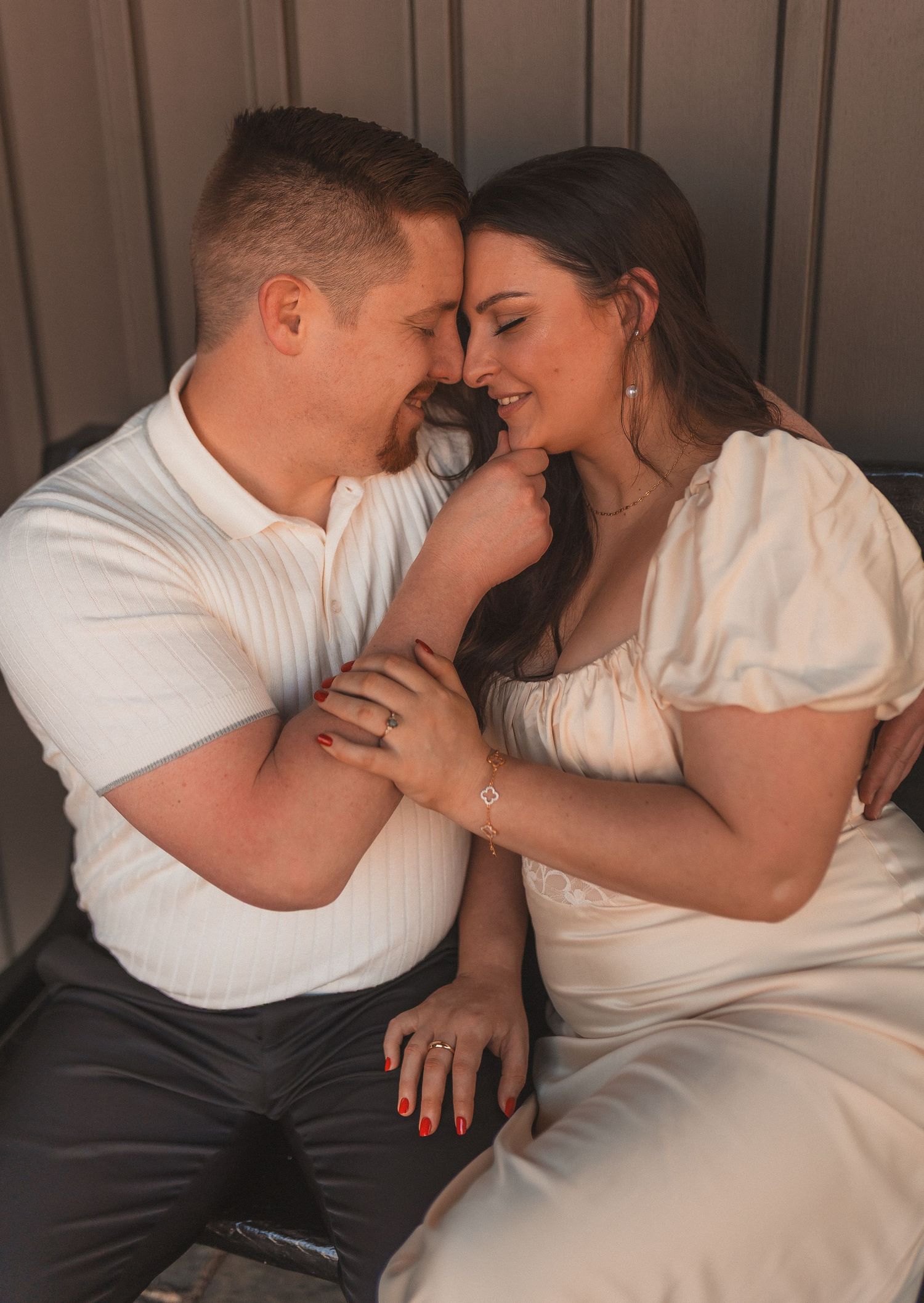 A romantic couple in white outfits shares an intimate moment while sitting close together in soft lighting.
