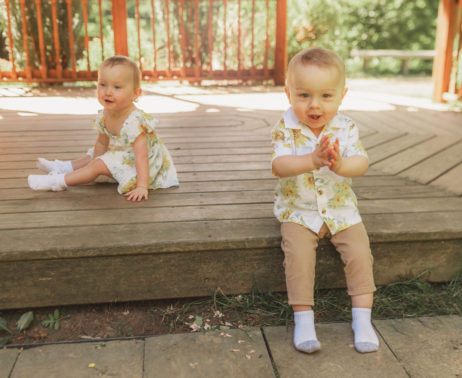Two toddlers in floral rompers and white socks sit and stand on a wooden deck on a sunny day.