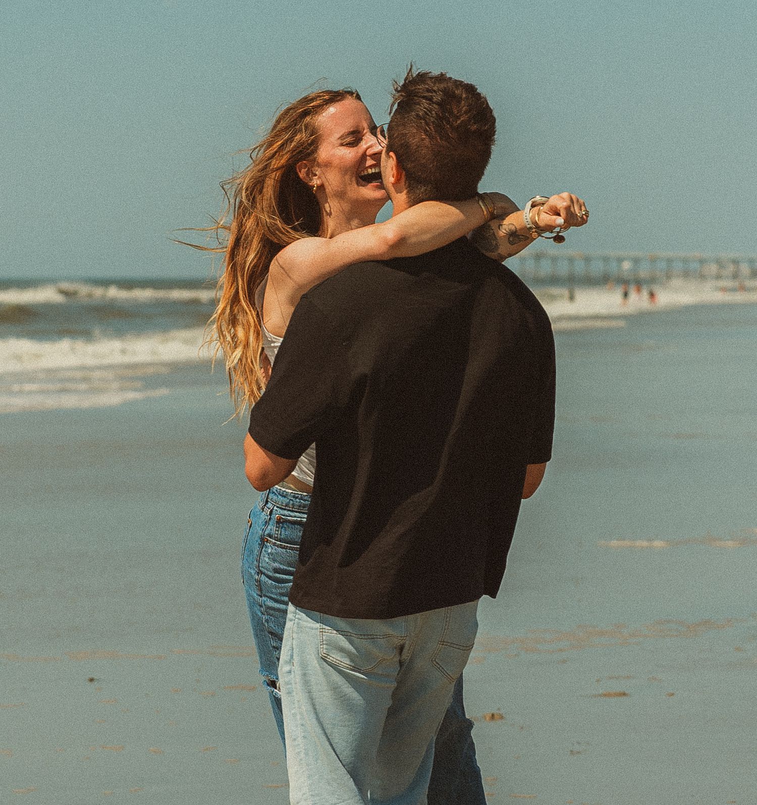 A couple sharing a romantic embrace and laugh together on a sunny beach with waves and pier in the background.