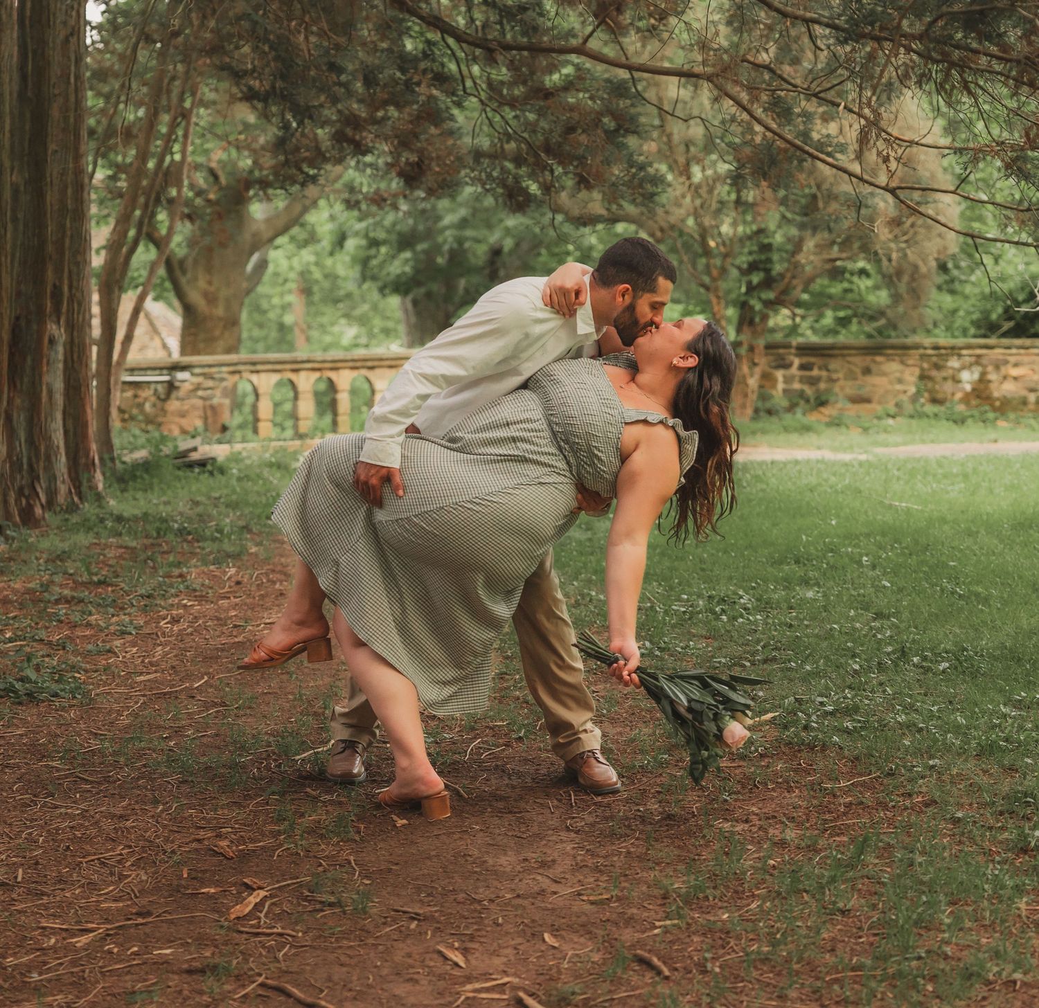 A romantic couple dancing in an intimate dip pose on a dirt path under evergreen trees in a park setting.
