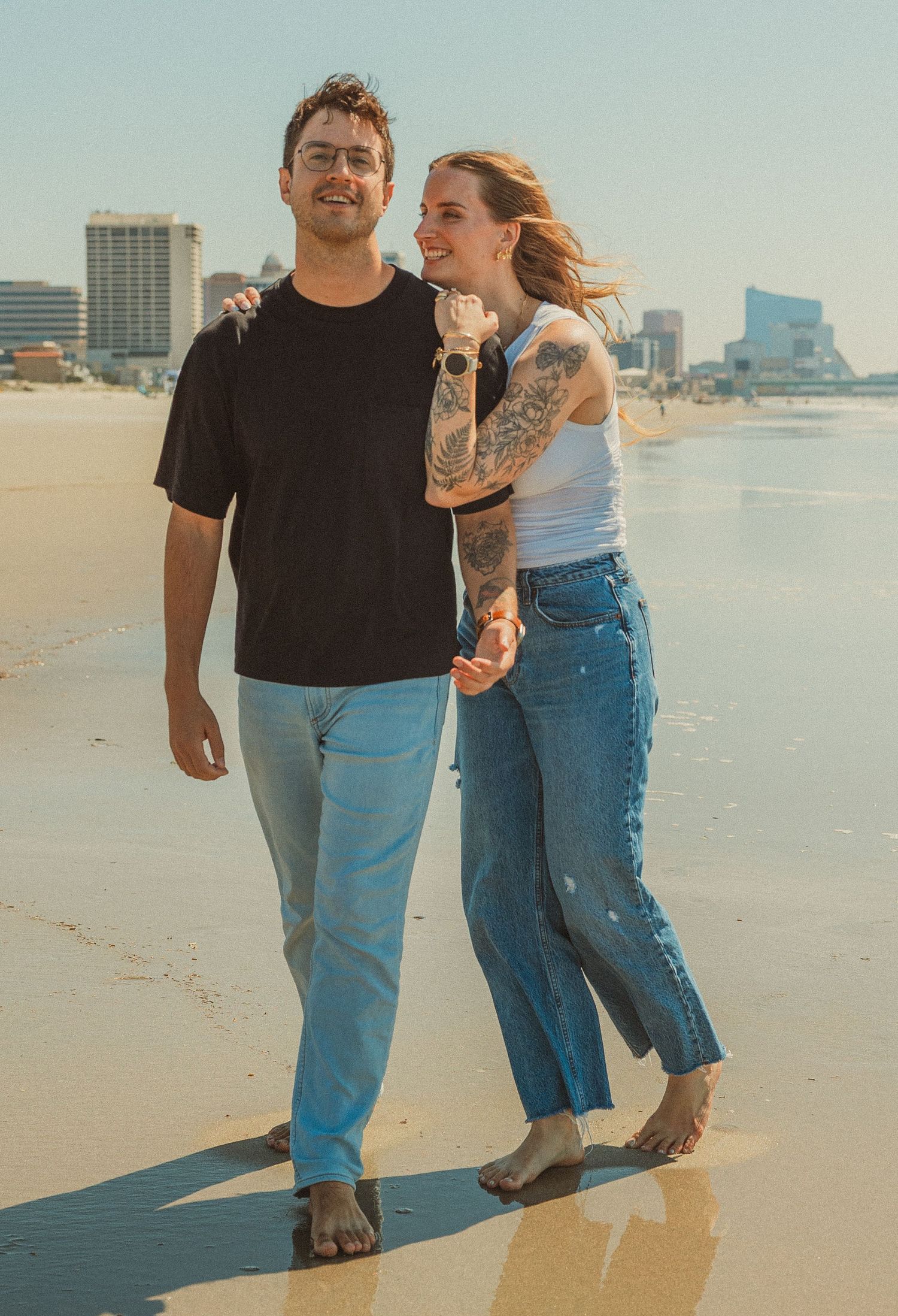 A couple embracing while walking barefoot on a sunny beach with city buildings visible in the background.