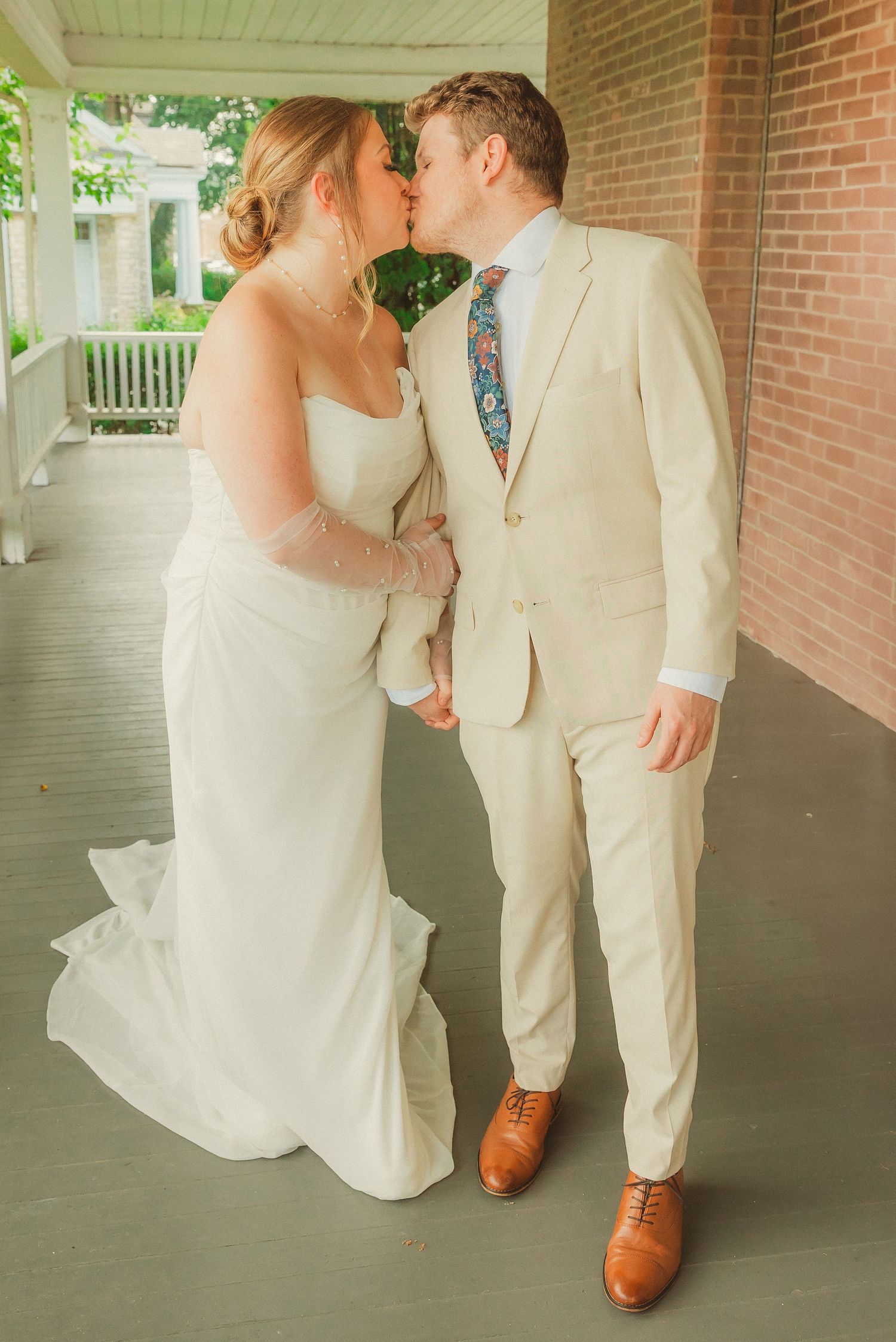 A romantic moment captured on a covered porch with the bride in white and groom in cream suit.