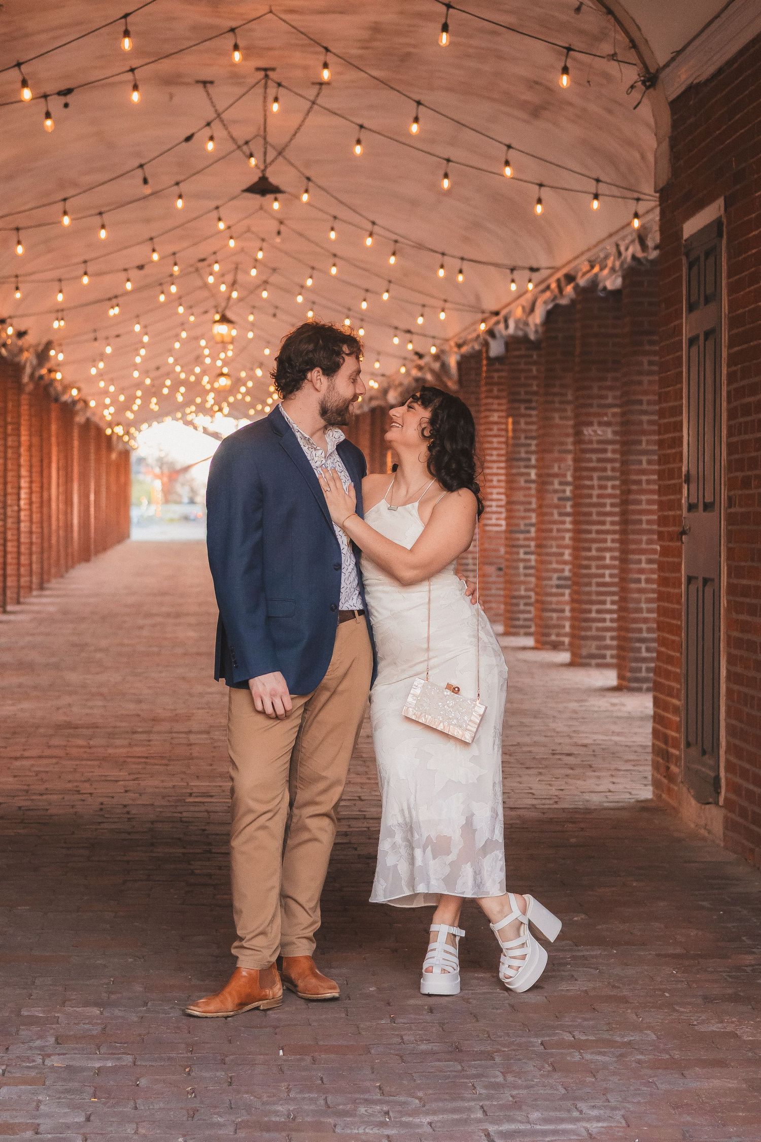 A couple poses under string lights in a brick archway corridor wearing formal attire.