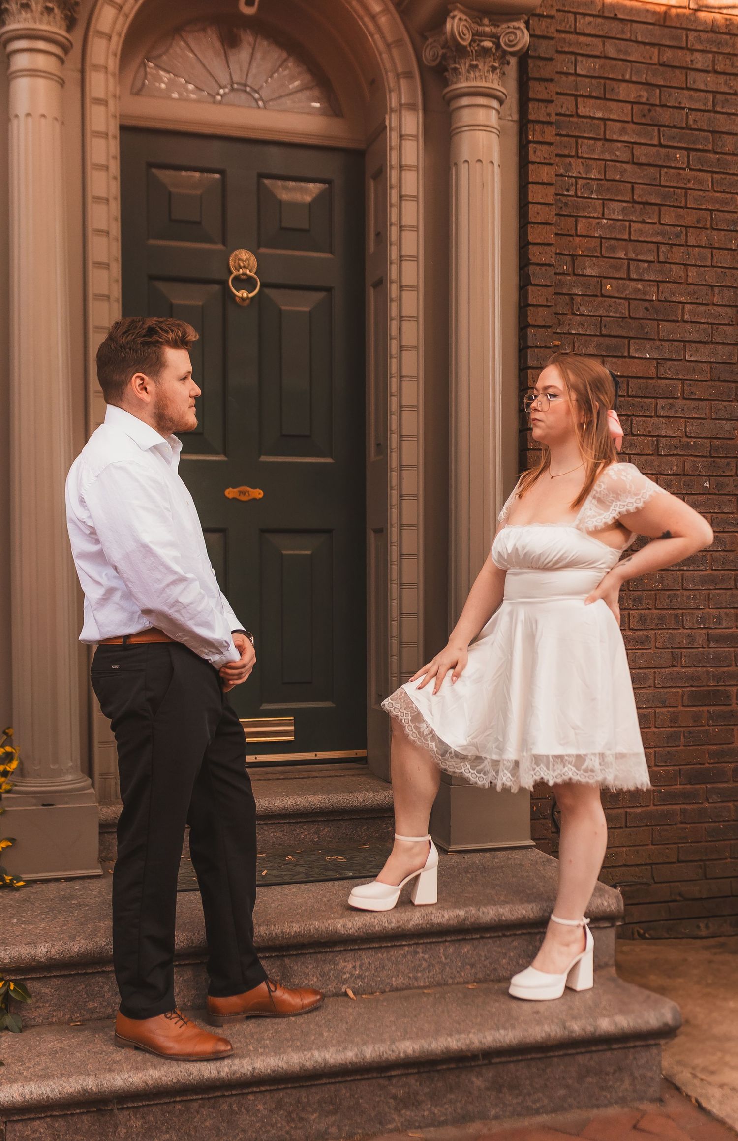 A couple in formal attire poses on brick steps in front of an elegant doorway.