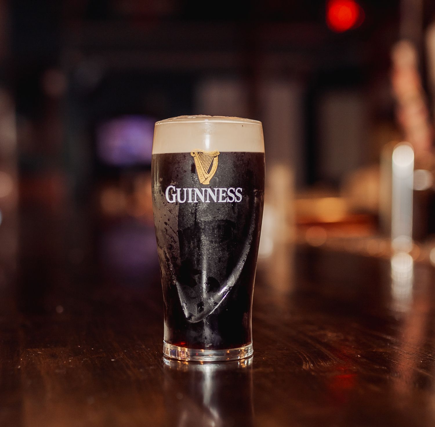 A pint glass of dark Guinness beer sits on a wooden bar with a creamy head and iconic logo visible in soft lighting.