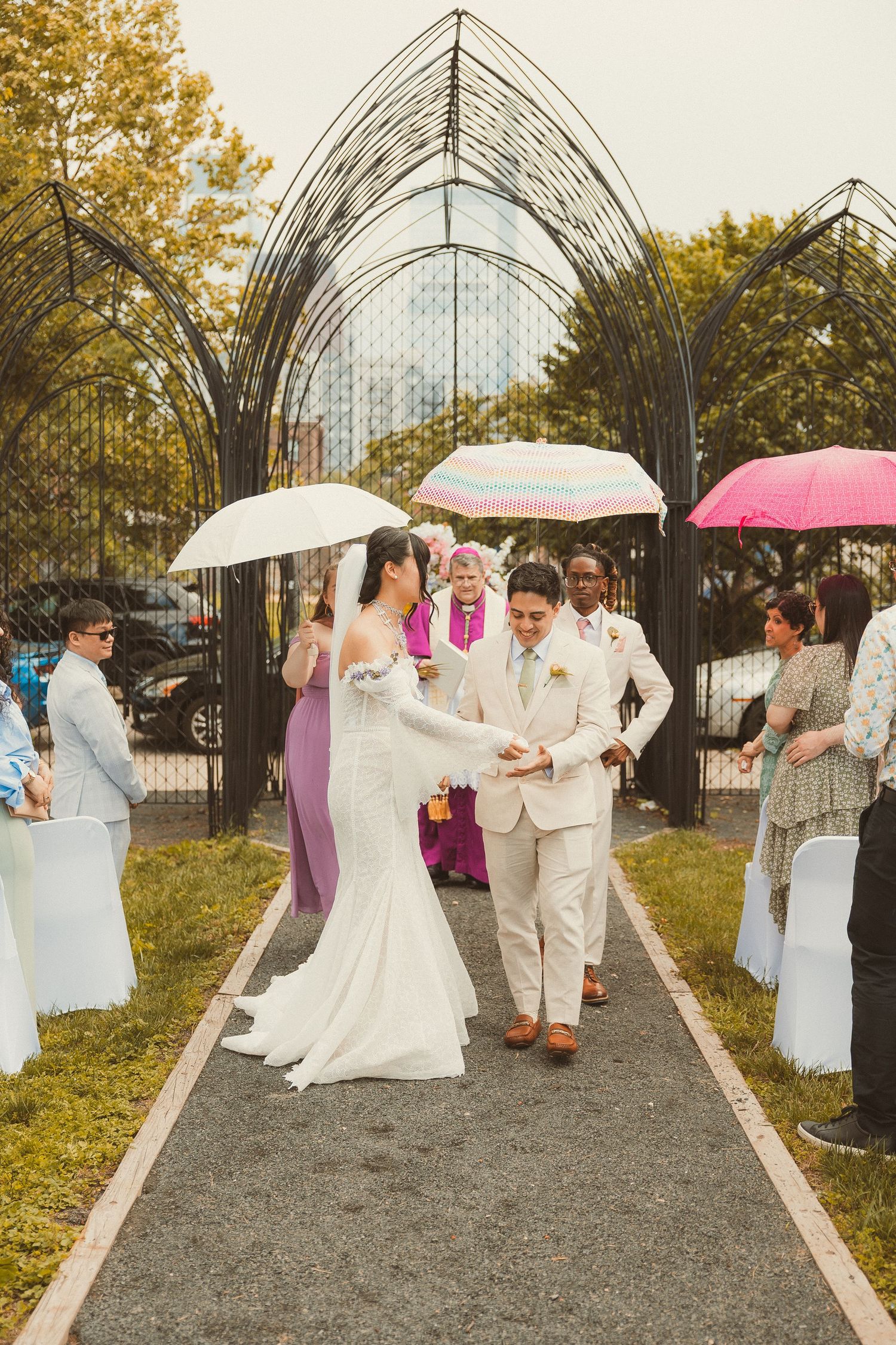 Wedding ceremony under an arched metal structure with guests holding umbrellas on a rainy day.