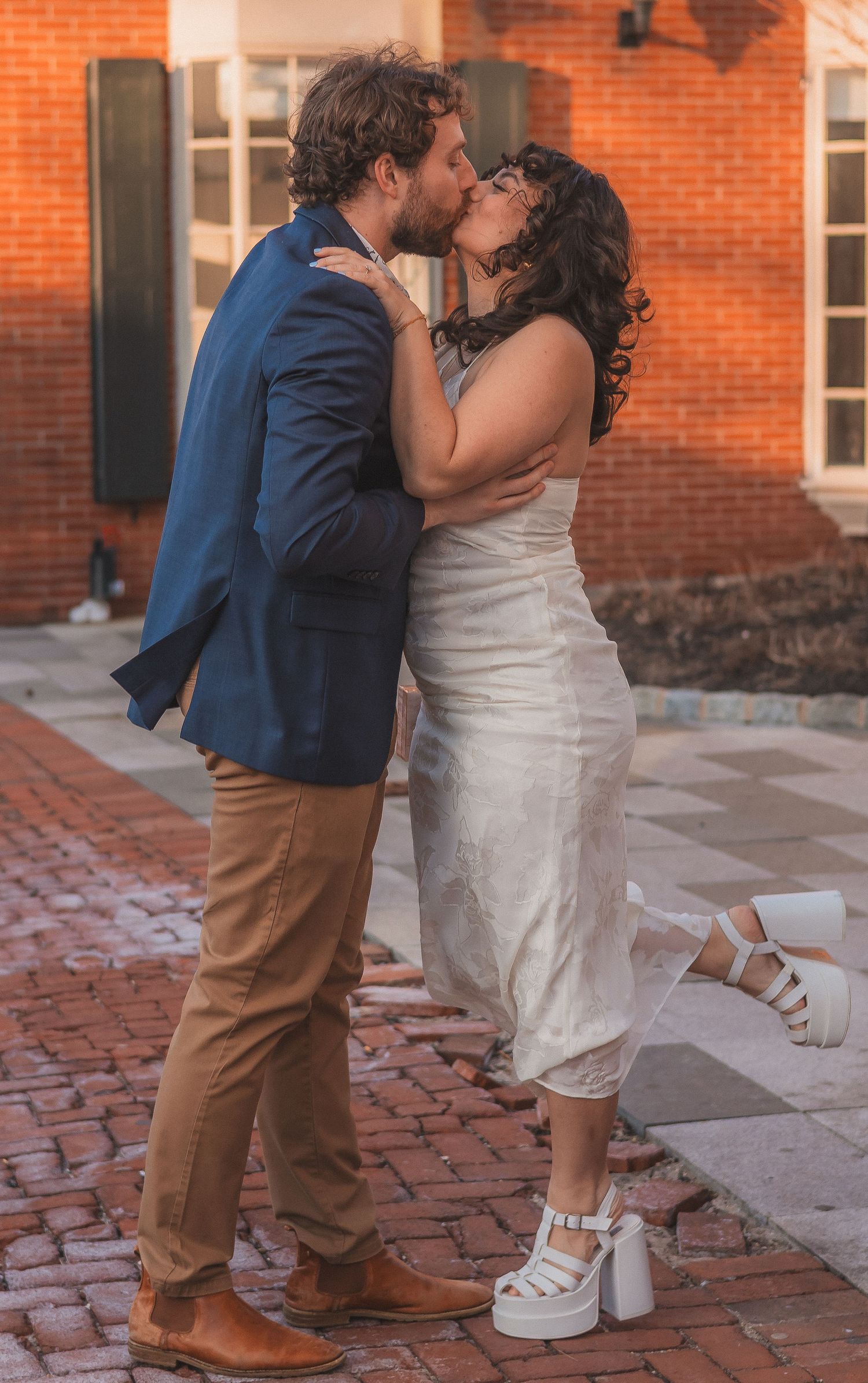 A couple embraces in a romantic kiss on a brick sidewalk.