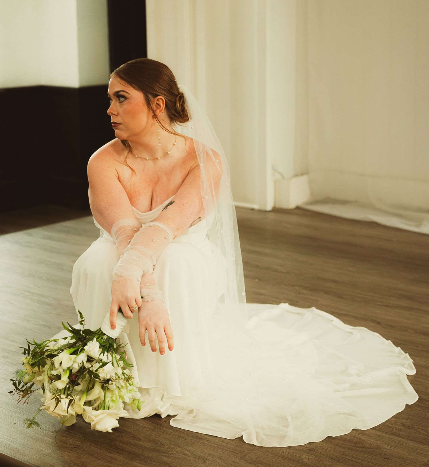 A bride in a flowing white gown and veil sits with a bouquet of white flowers and greenery beside her on the floor.