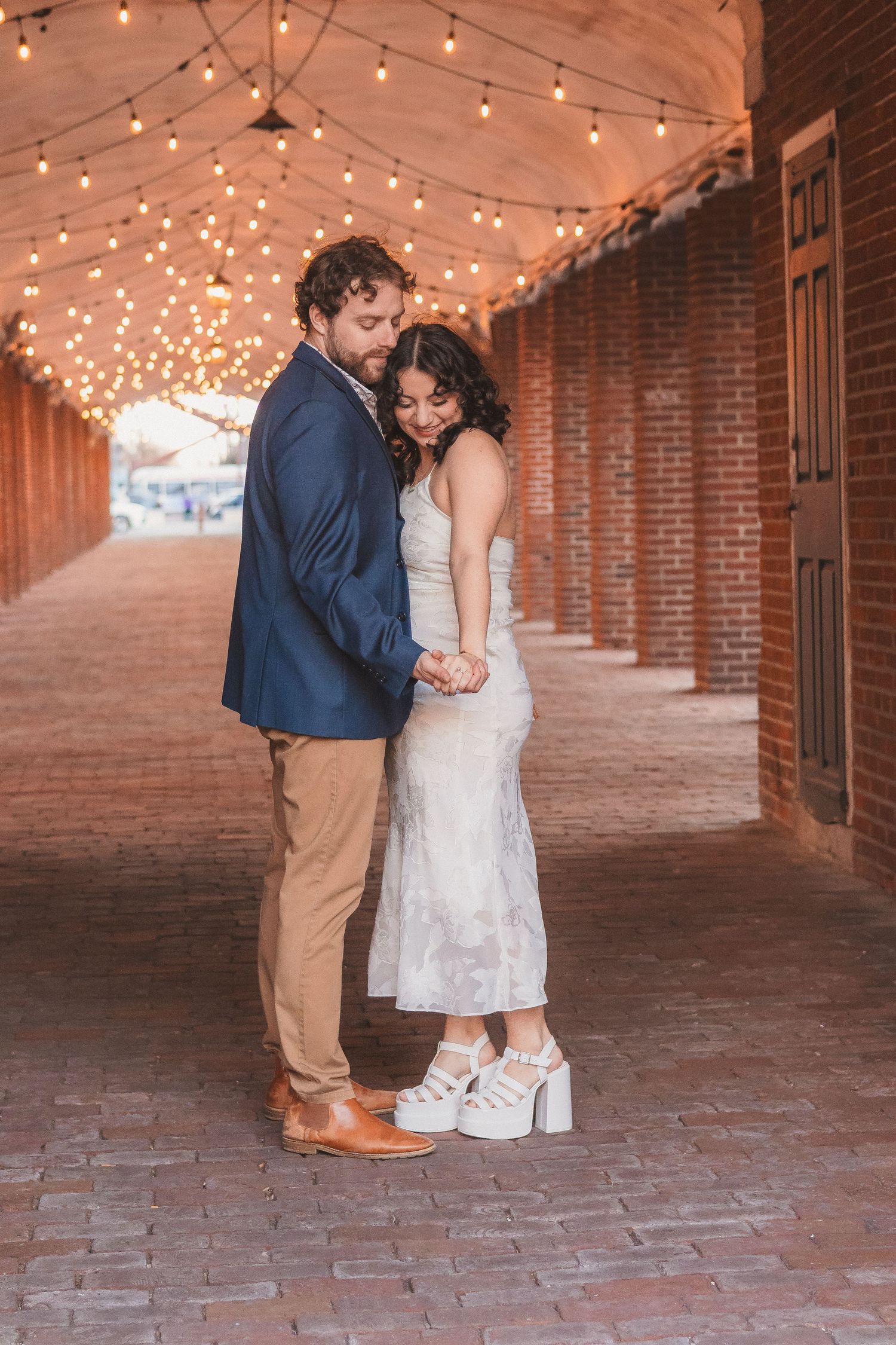 Romantic couple embracing in formal wear under twinkling lights in a brick passageway.