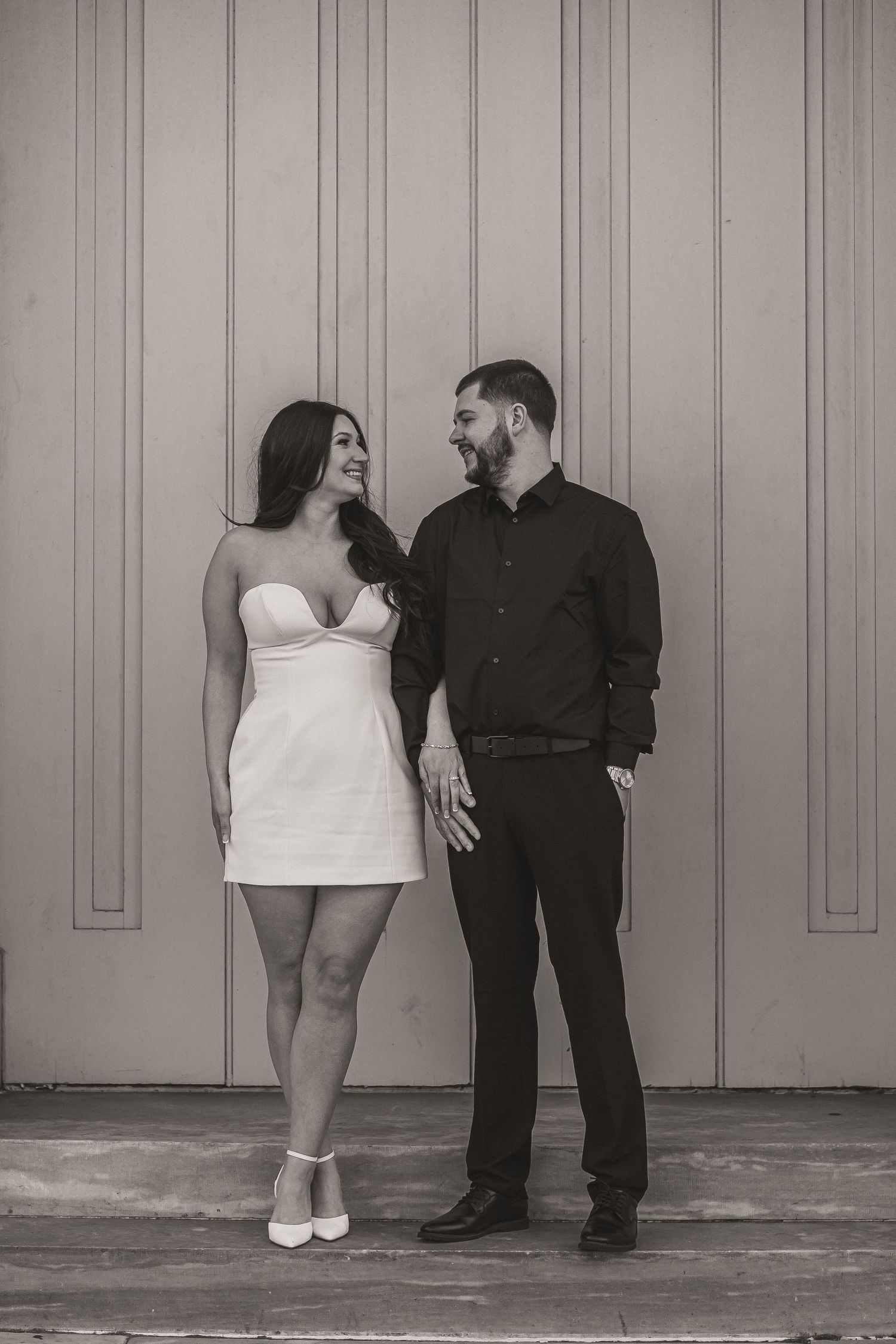 A couple in formal attire pose together against a grey wall in black and white.