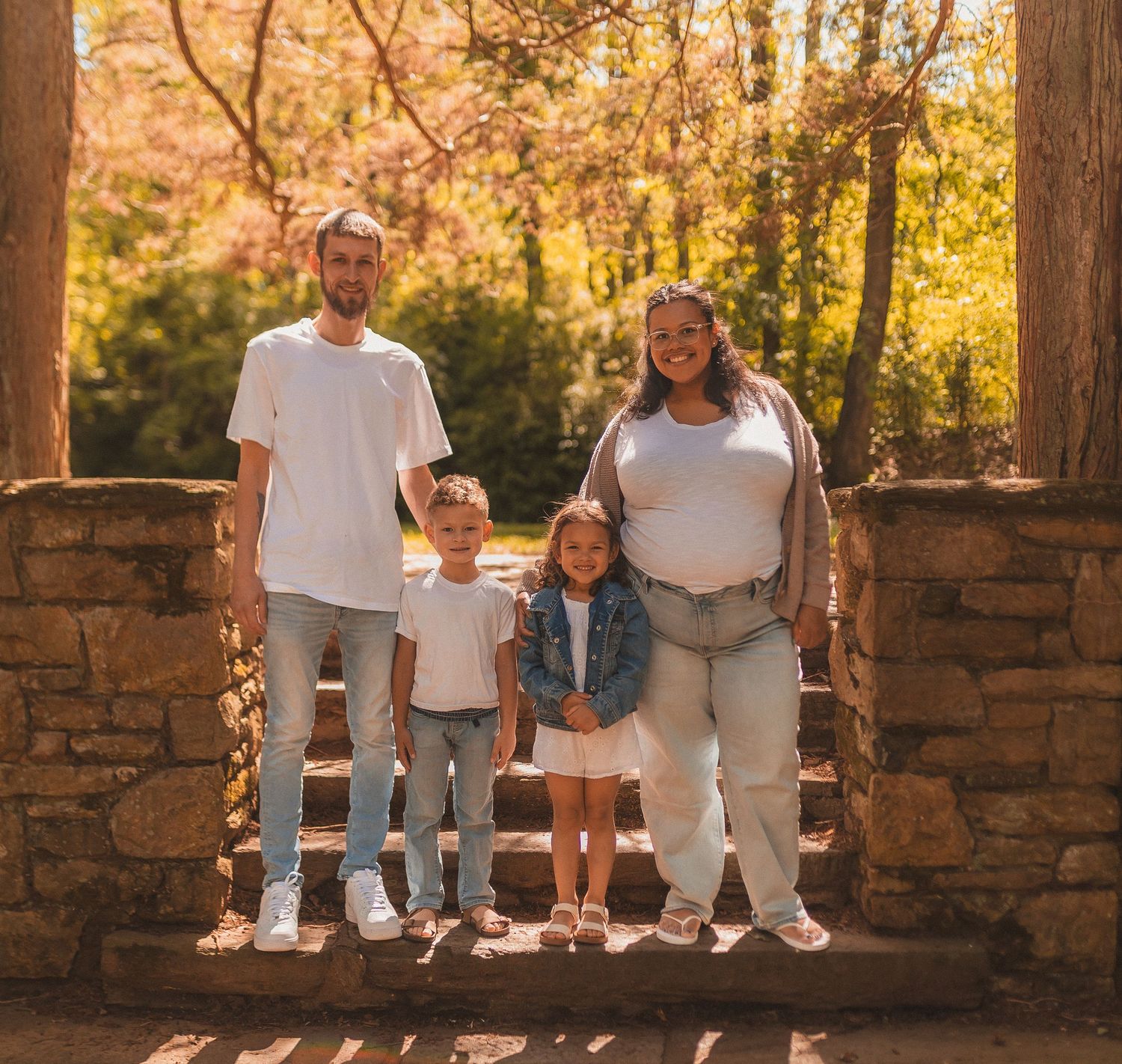 A family of four stands together on stone steps surrounded by golden autumn foliage in warm sunlight.