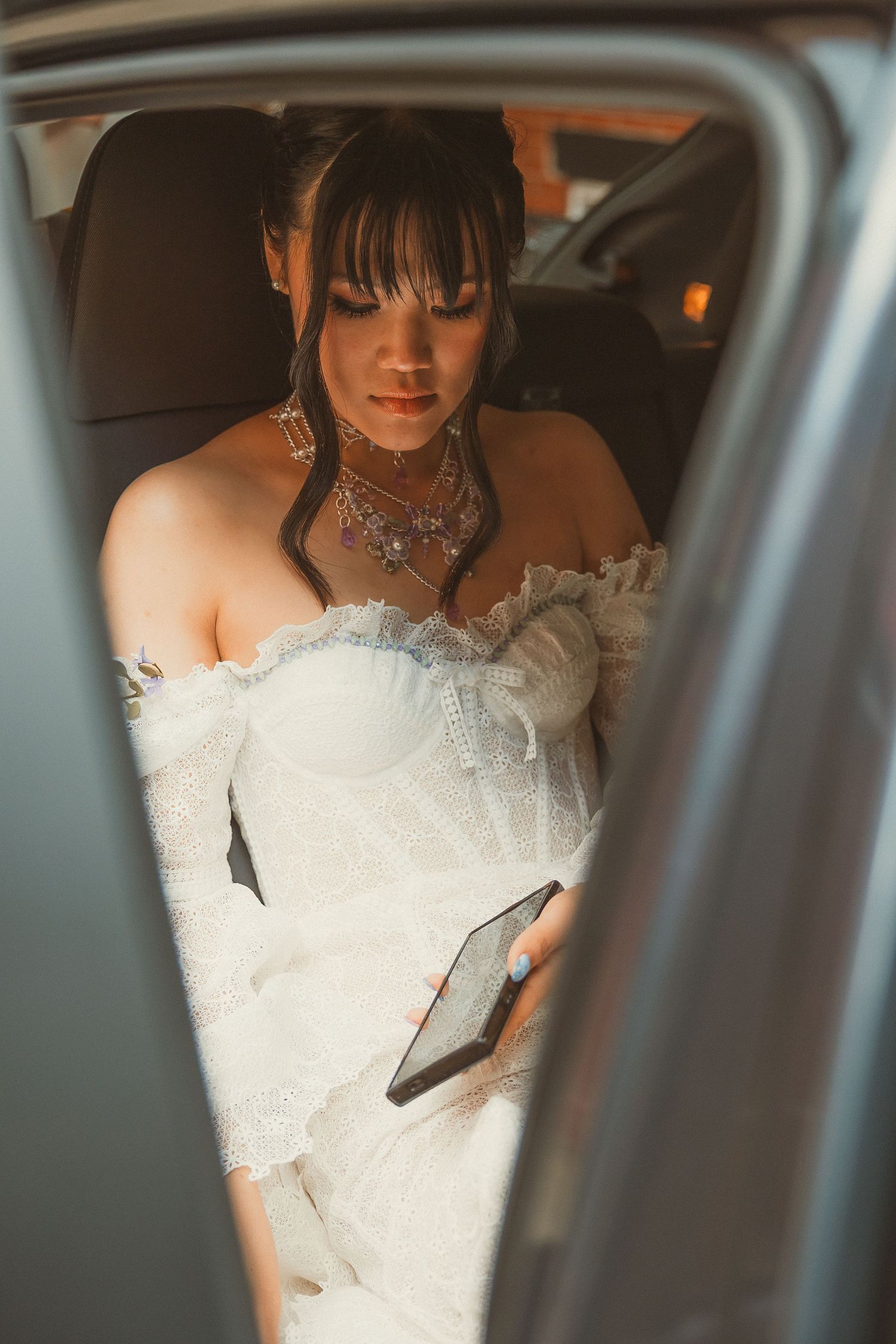 Close-up shot through a car window showing an elegant white wedding dress and statement necklace.