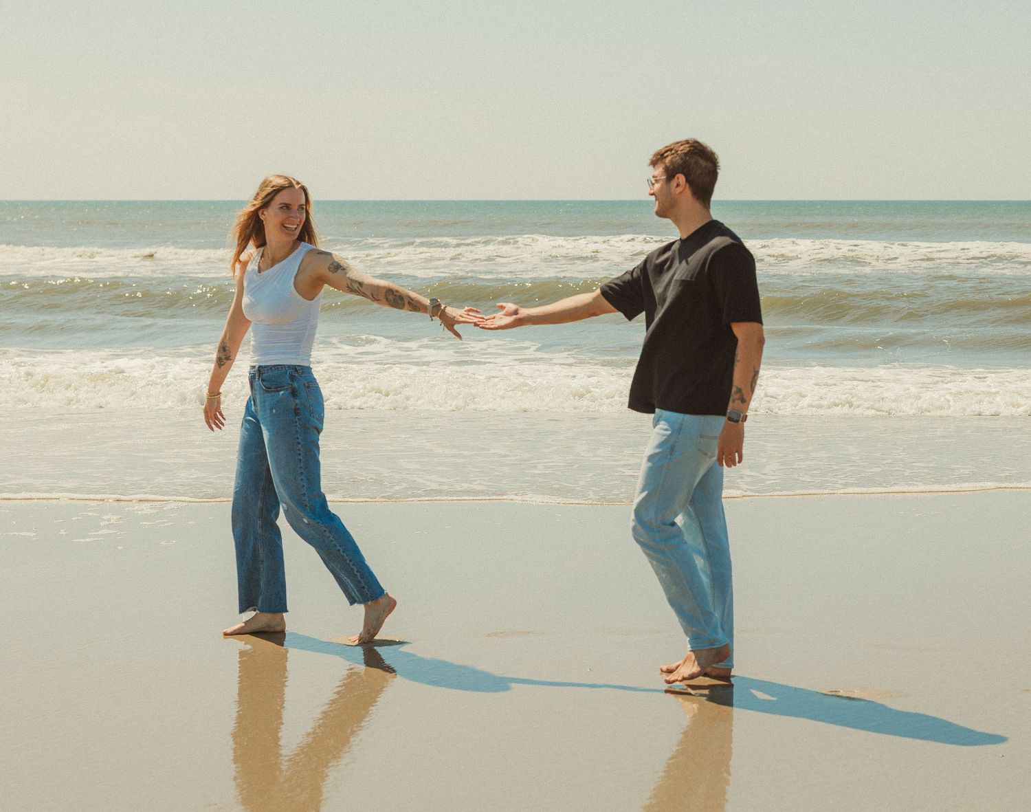 Two people hold hands while walking along a sunny beach with waves and ocean in the background.