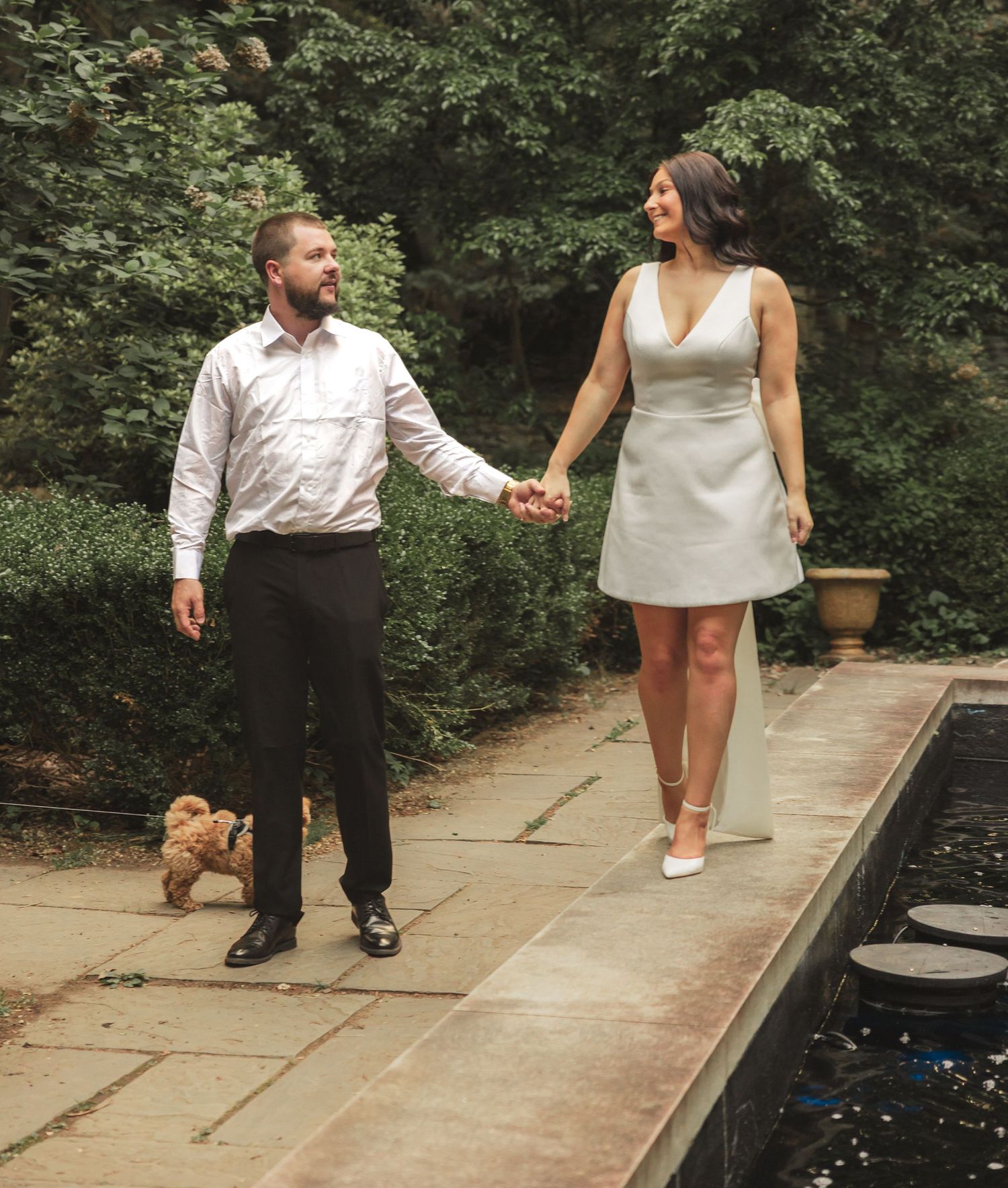 A couple in formal attire walks along a stone path beside a water feature in a garden setting.
