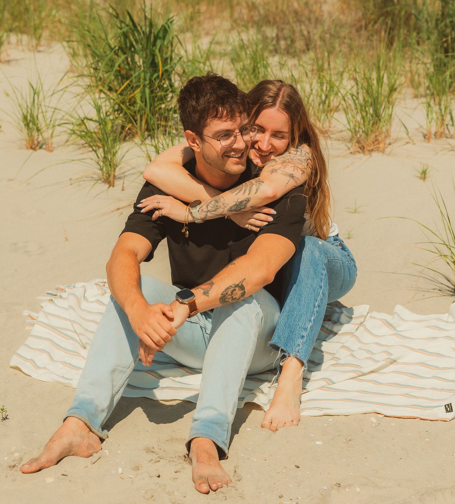Two affectionate people cuddling and laughing together while sitting on a striped blanket at the beach.