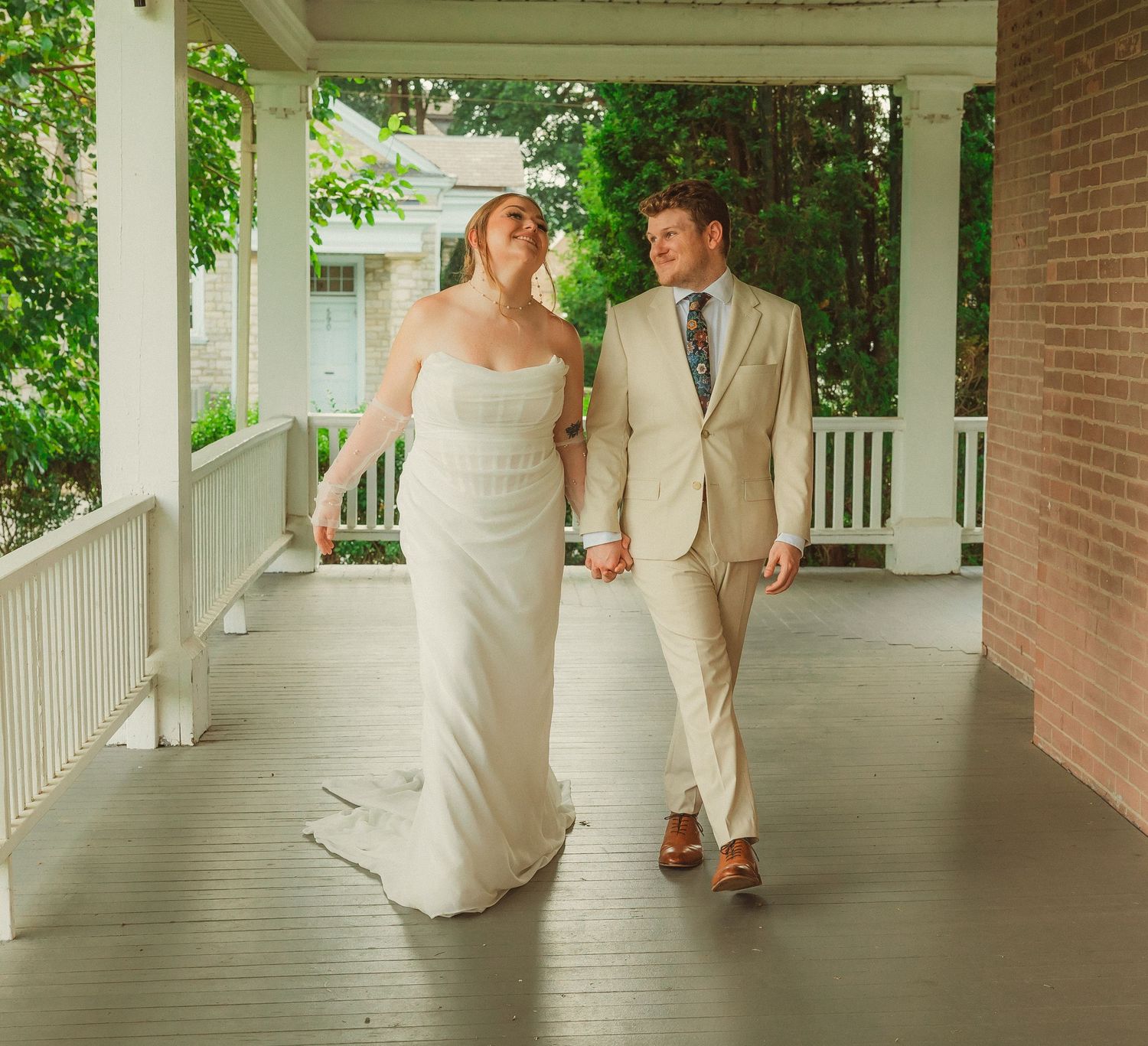 A couple walks hand in hand along a covered porch with white columns and brick walls.