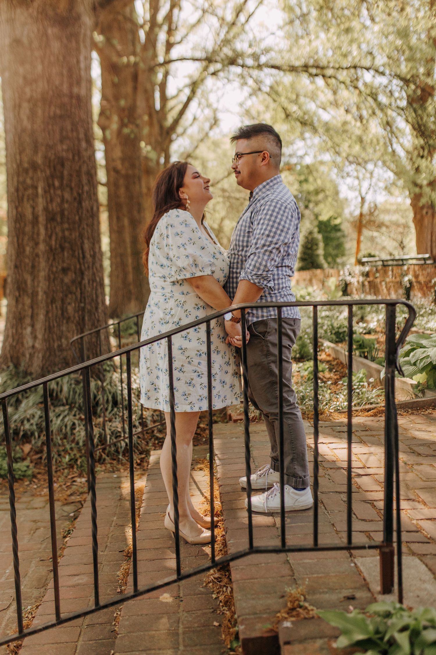 A couple shares a loving moment on brick steps with iron railings in a wooded garden setting.