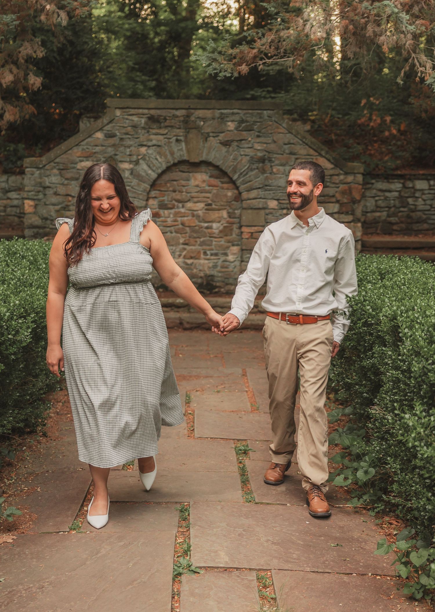 A couple walks hand in hand along a stone garden path with an arched stone wall and greenery in the background.