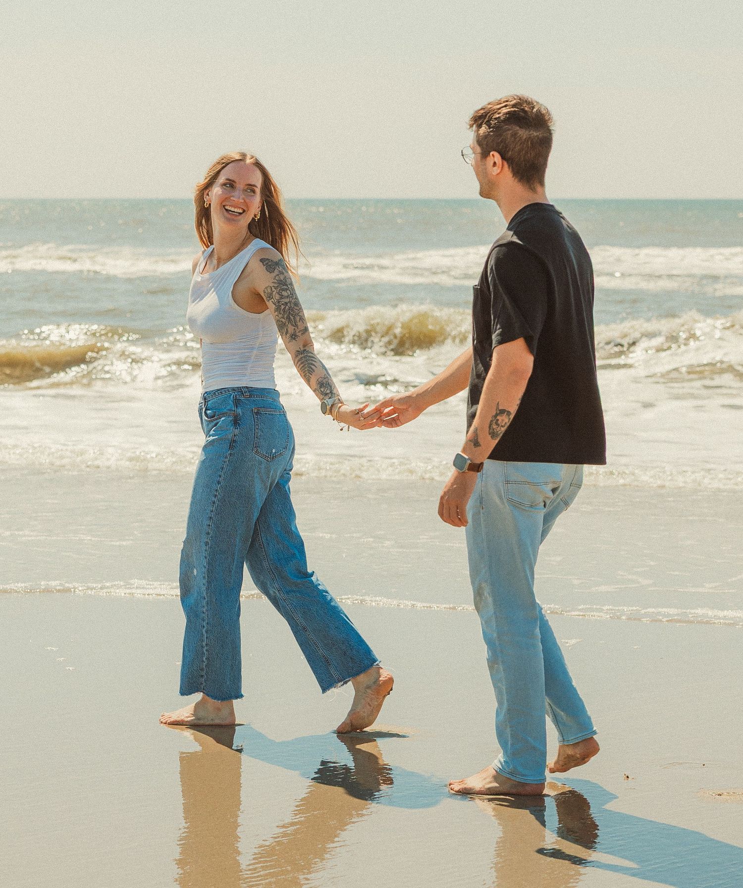Two people hold hands while splashing together in the shallow ocean waves on a sunny beach.