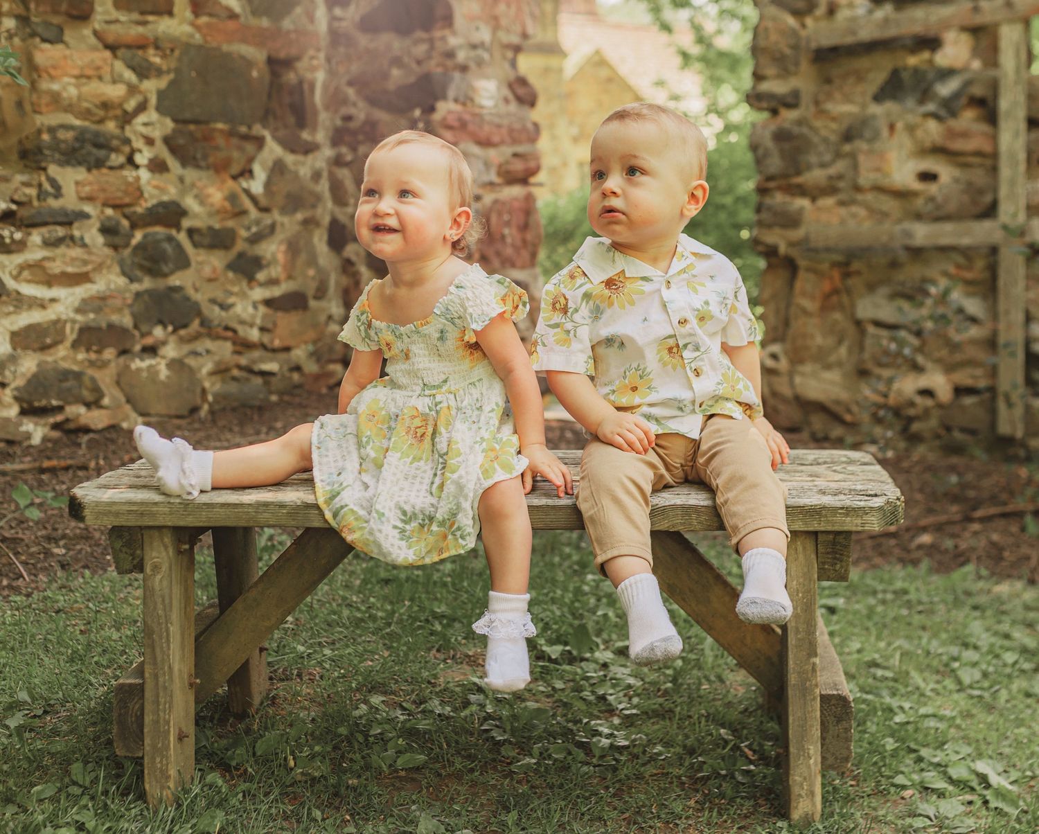 Two toddlers laugh and interact while sitting together on a wooden bench in a rustic garden setting.