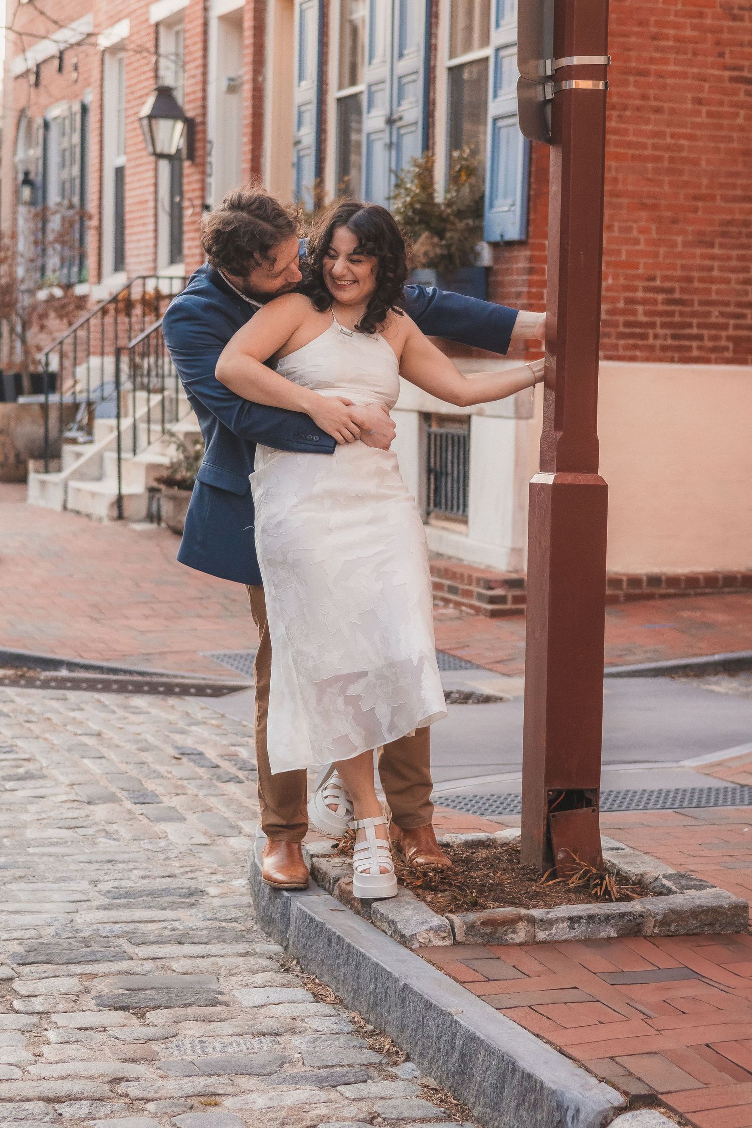 A couple shares a playful moment near a lamp post on a brick sidewalk.