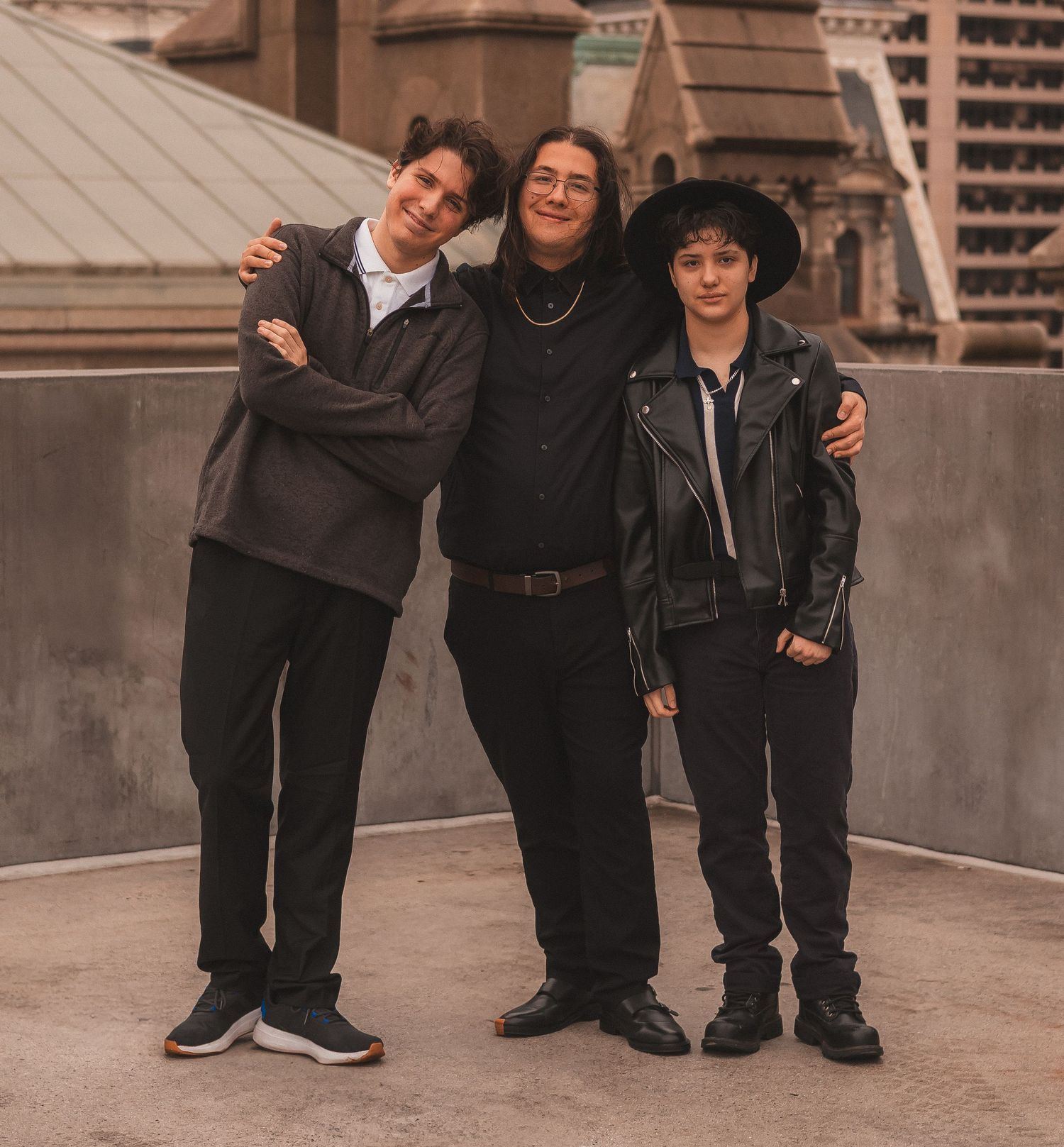Three friends in dark clothing embrace while sharing a joyful moment outdoors.