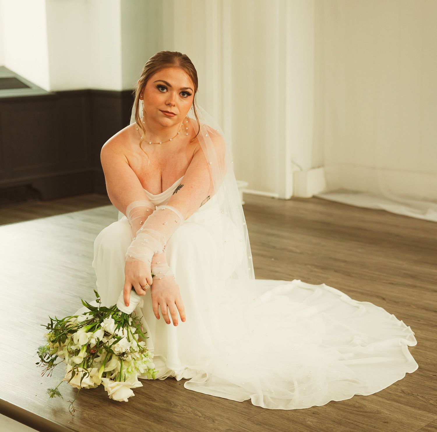 A bridal portrait in white gown with flowing veil sits on hardwood floor holding a delicate floral bouquet.