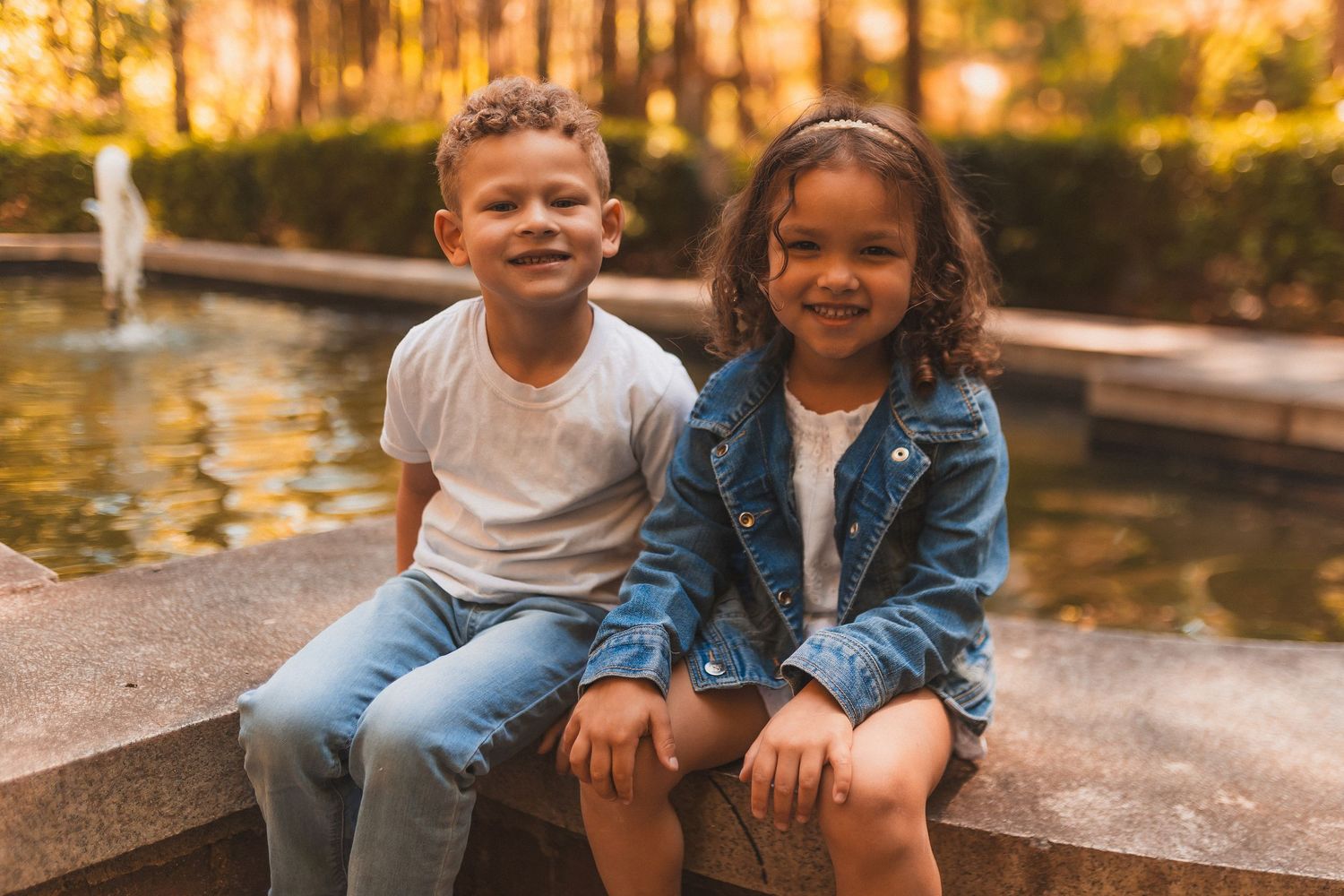 Two children sit together by a fountain at sunset in a park, smiling warmly in casual outfits.