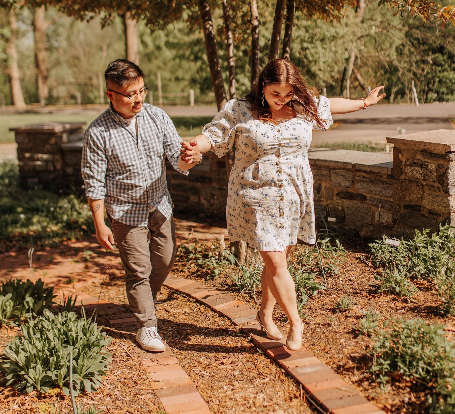 A couple walks hand in hand along a stone path in a garden on a sunny afternoon.