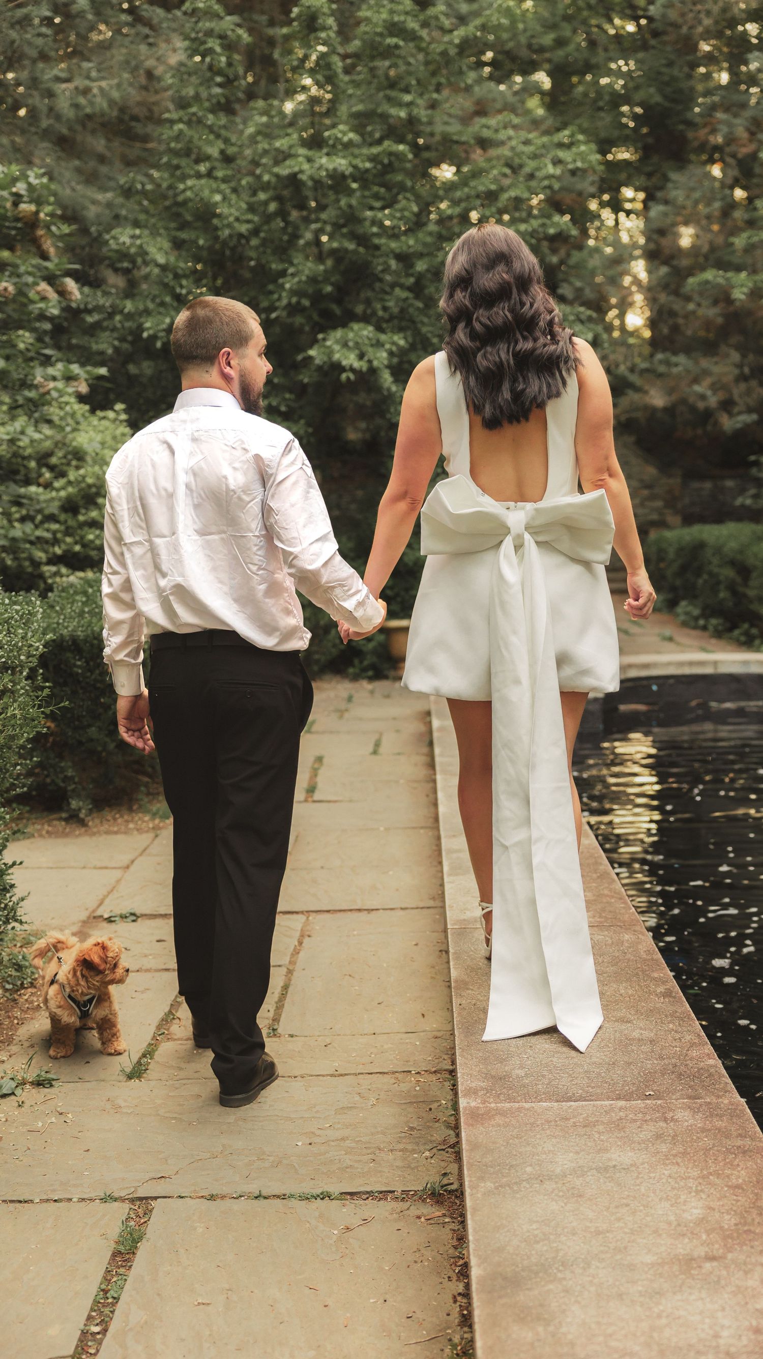 Couple walking along a pond path with their small dog, bride wearing a white dress with large back bow detail.