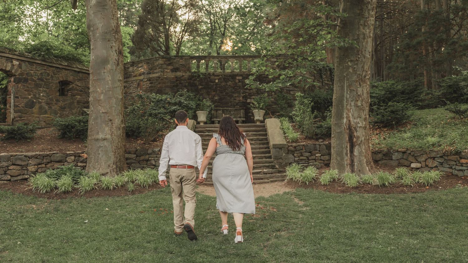 Tree-lined pathway in a lush wooded area with stone walls and steps leading up a gentle slope.