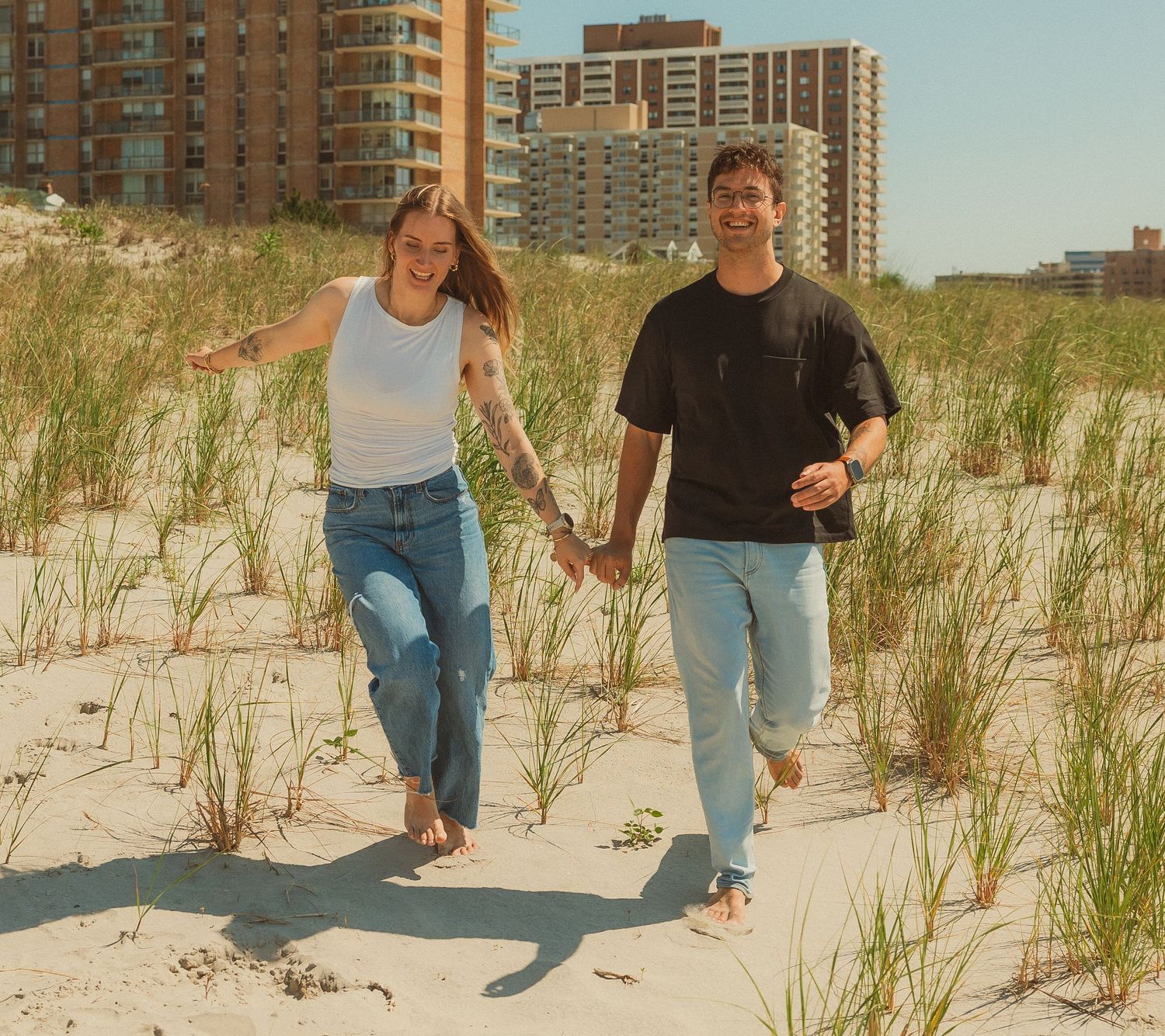 People walking along a sandy beach path with coastal grass and tall apartment buildings visible in the background.