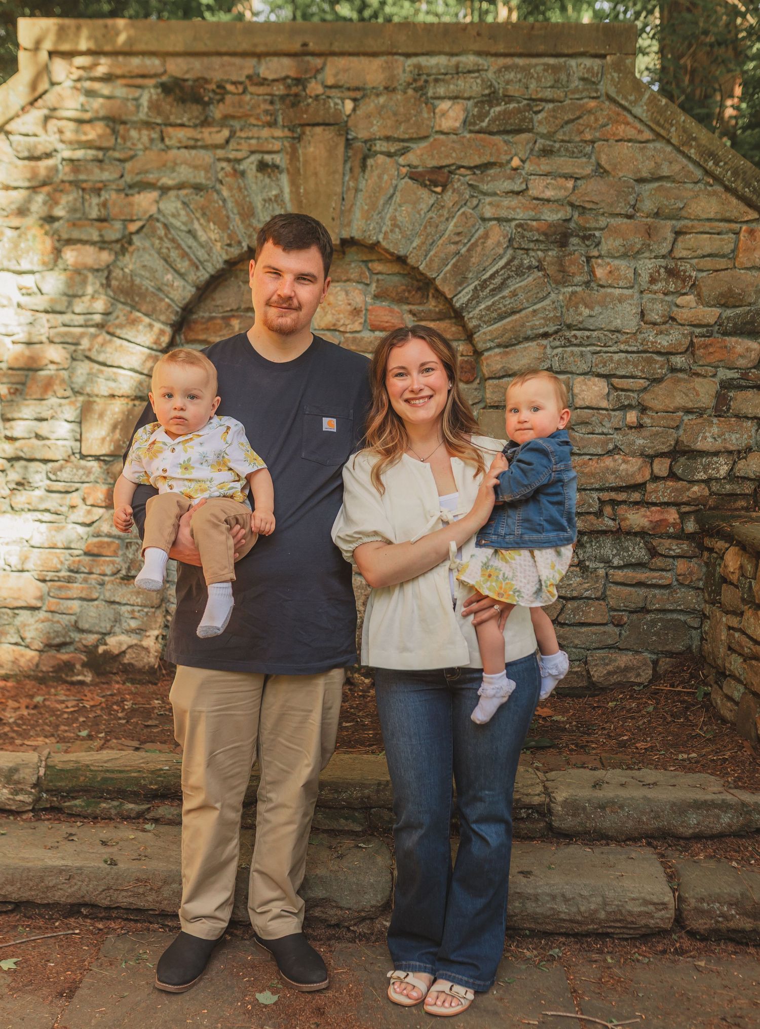 A family of four stands together in front of a rustic stone archway outdoors during a photo session.
