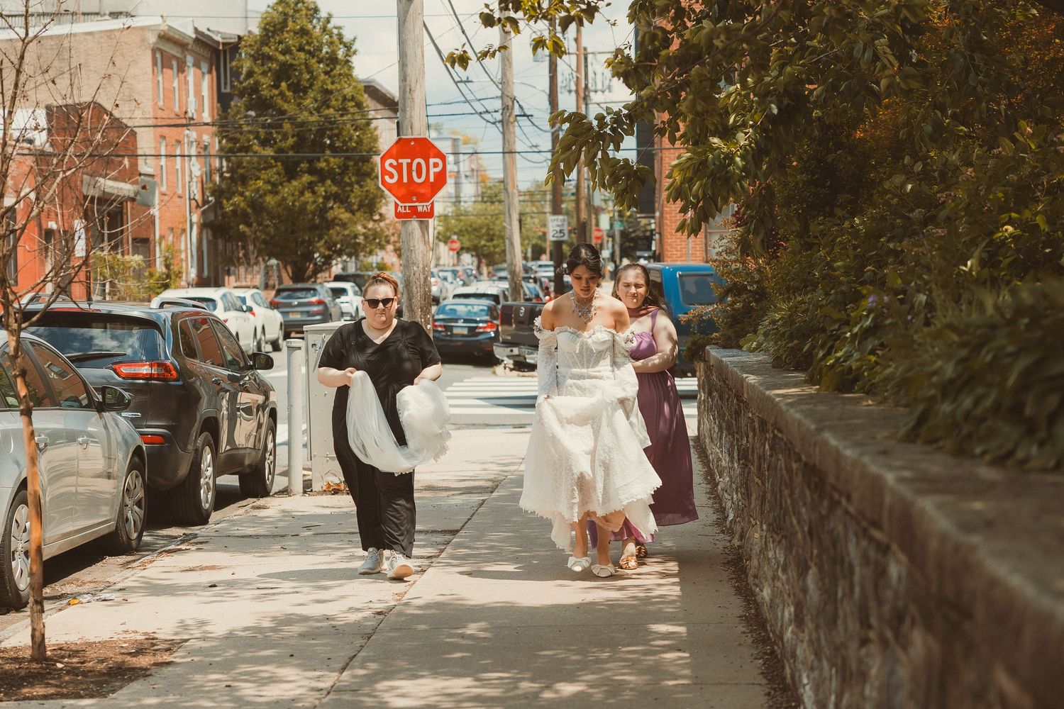 A couple walks along a stone wall-lined sidewalk in an urban neighborhood on a sunny day.