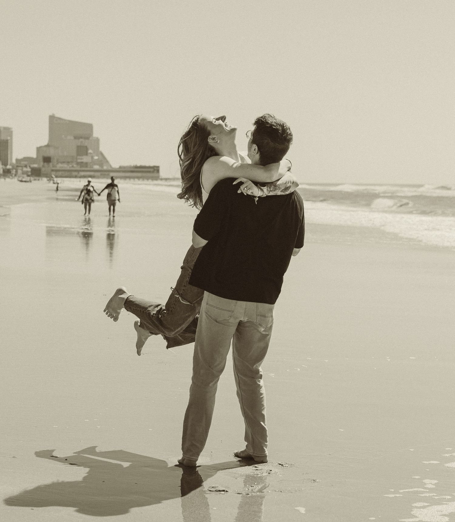 A romantic black and white scene of a couple embracing on a beach with silhouettes of people walking in the background.