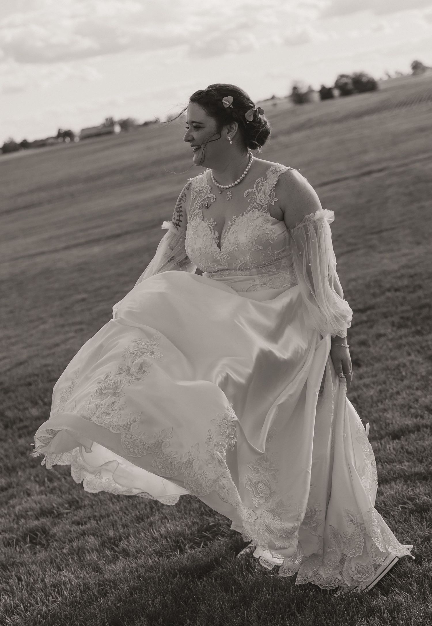 Elegant vintage-style wedding dress with intricate lace detail flows in the breeze across a countryside field in black and white.
