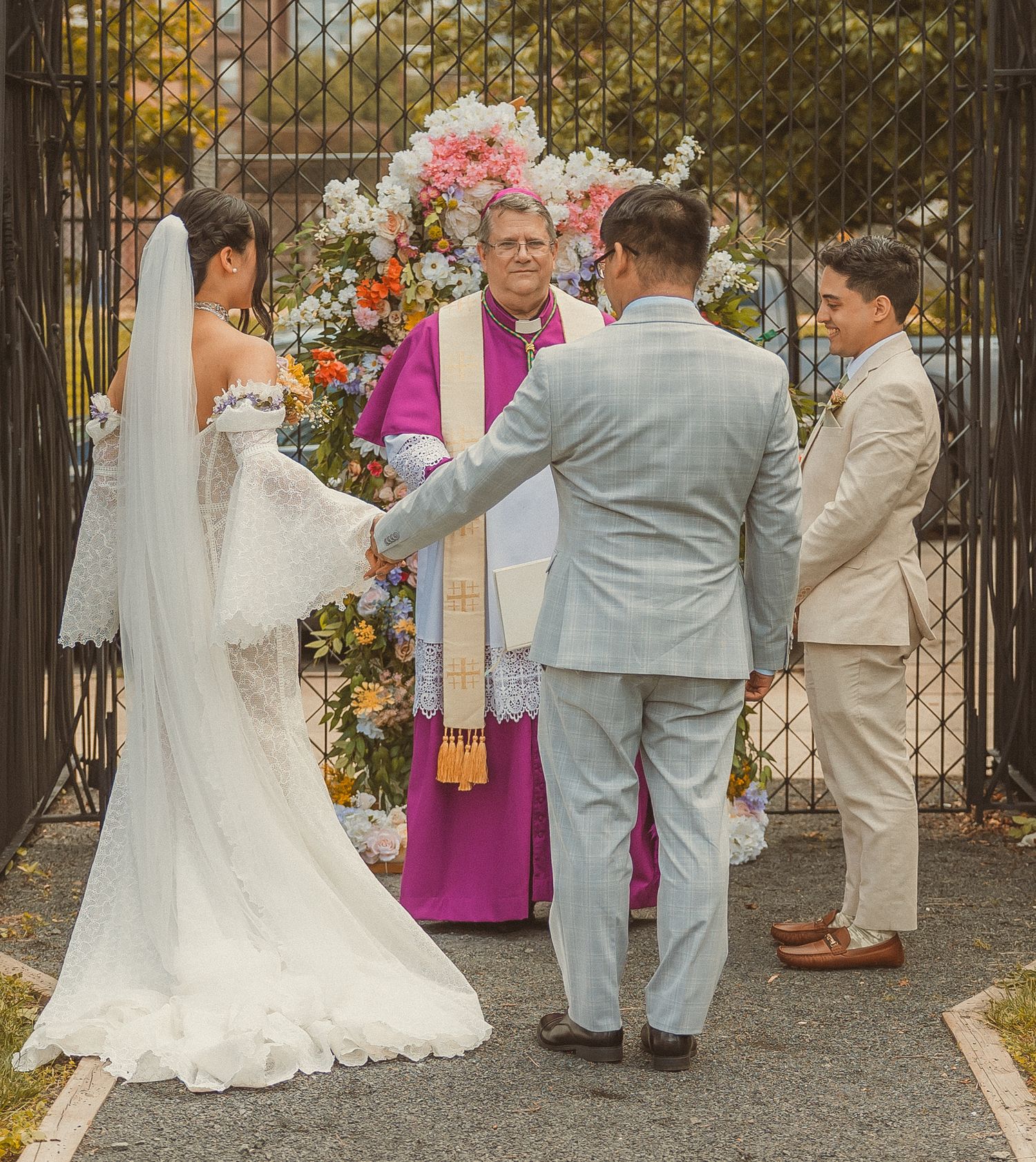 A wedding ceremony takes place under a floral arch with an officiant in pink robes presiding over the service.