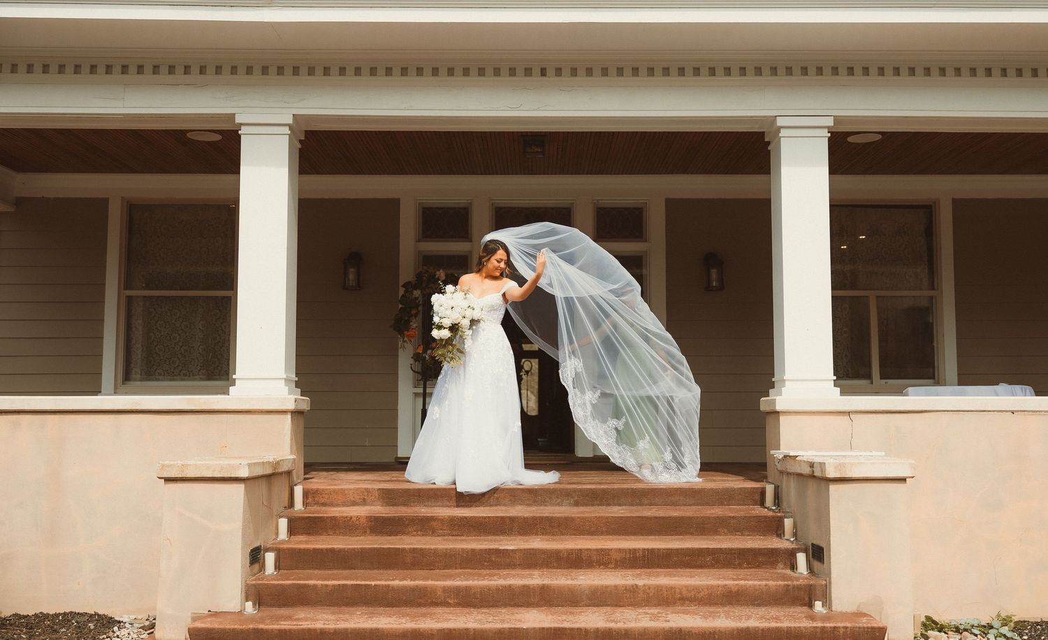 Wedding moment captured on cream-colored building steps with flowing veil in breeze.