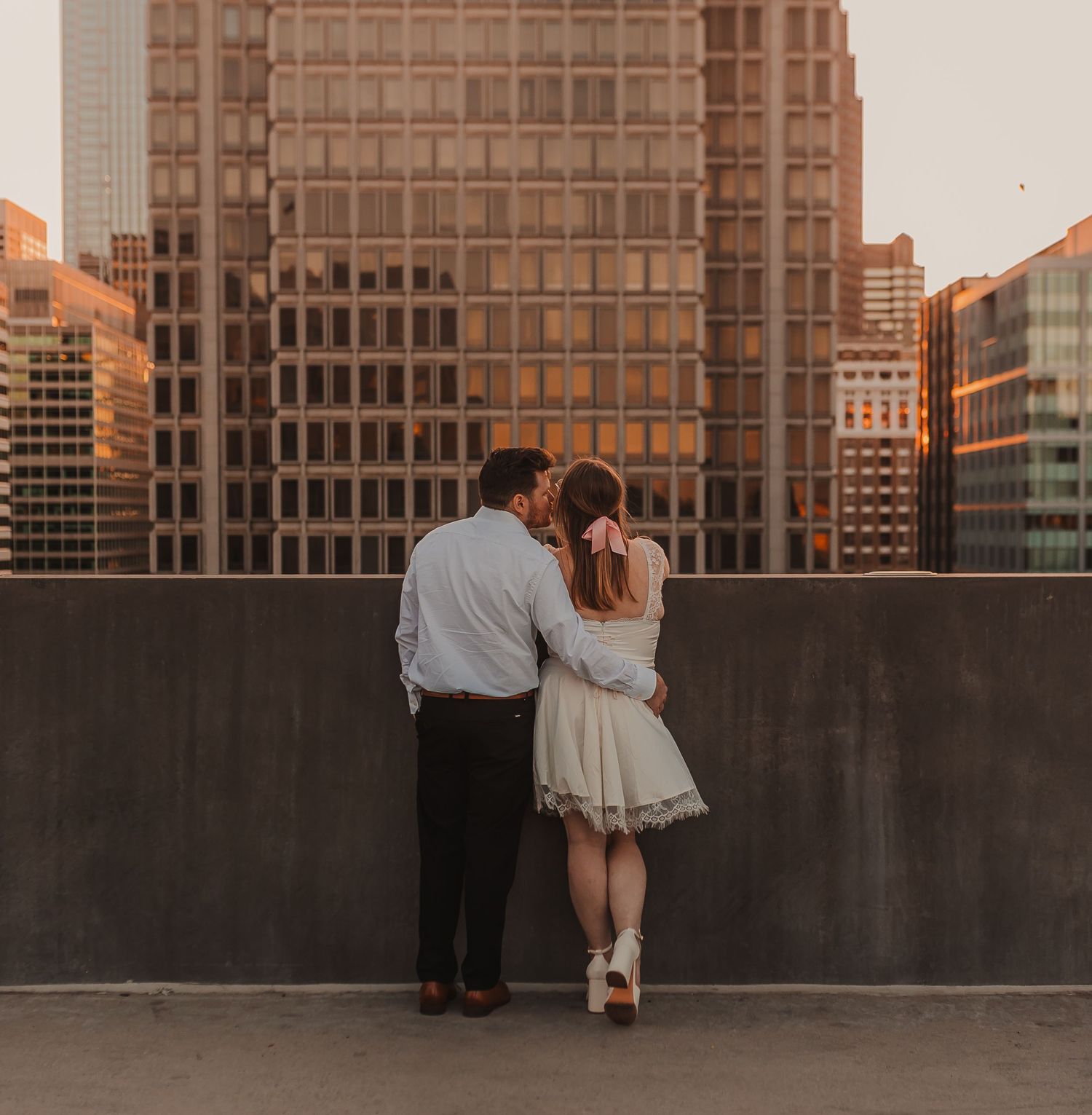 A romantic couple embraces on a rooftop at sunset with city buildings in the background.