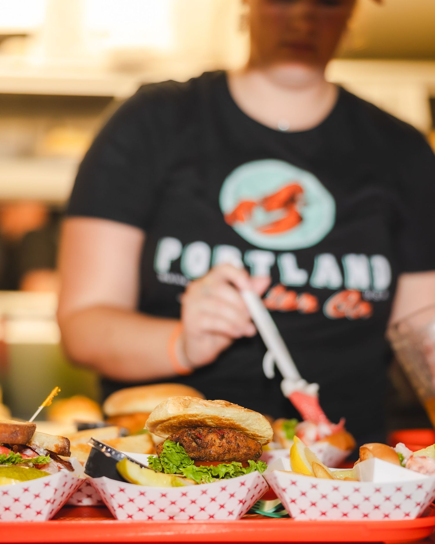 Portland lobster co. staff member serving gourmet burgers in red checkered food baskets at a food stand.