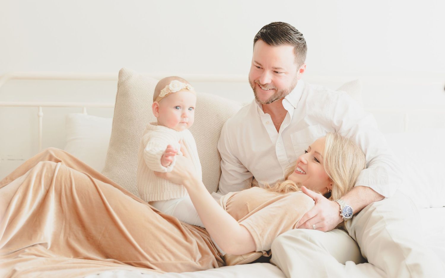 Happy family with a baby girl resting together on a cozy white bed in a bright, airy room.