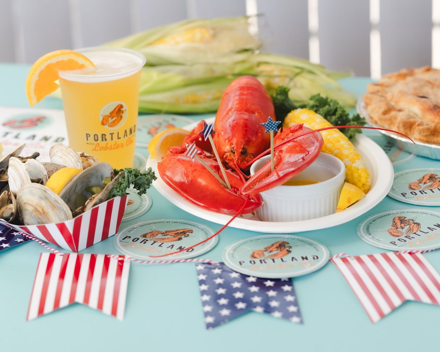 A festive Portland seafood spread with lobster, corn, clams, pie, and a beer on a patriotic decorated table.