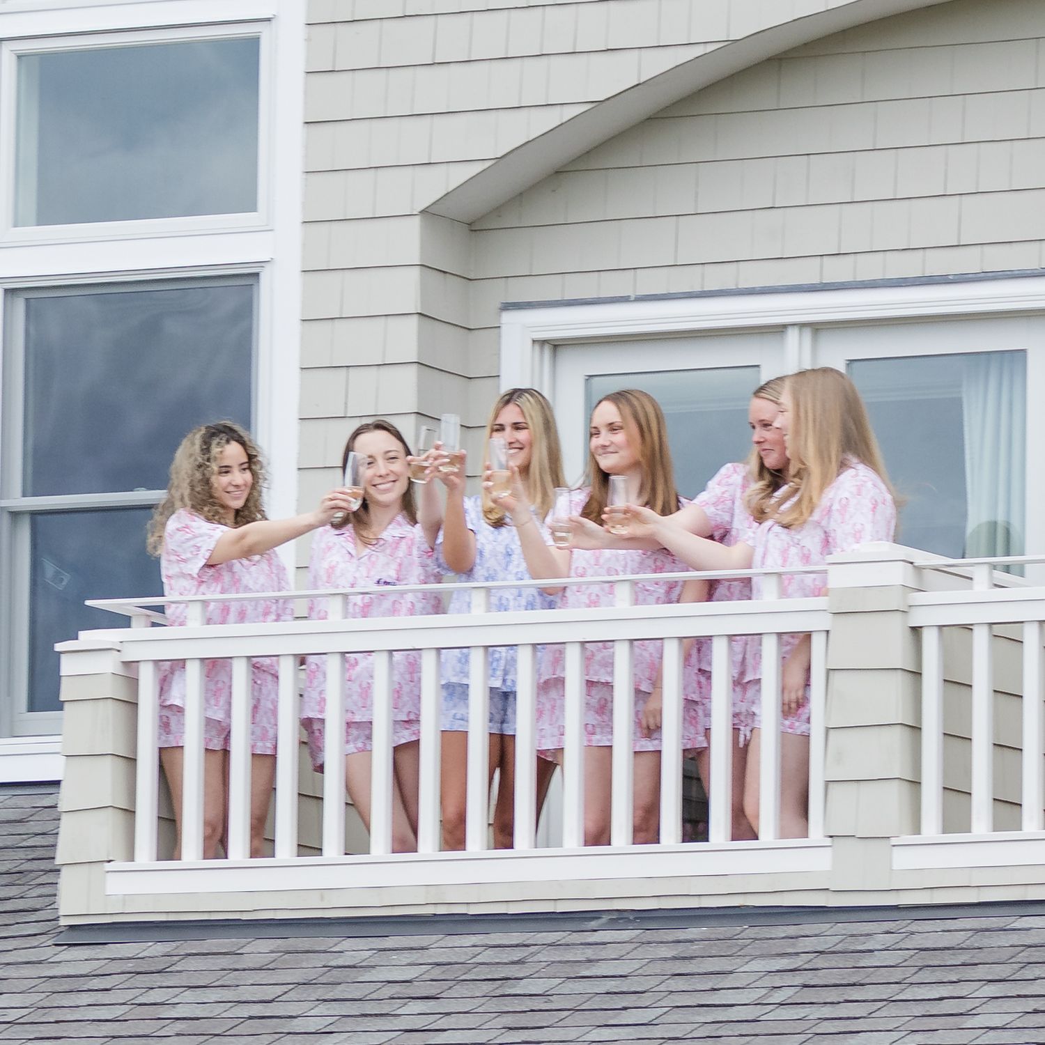 Five women in matching pink floral robes toast on a white balcony of a gray shingled beach house.