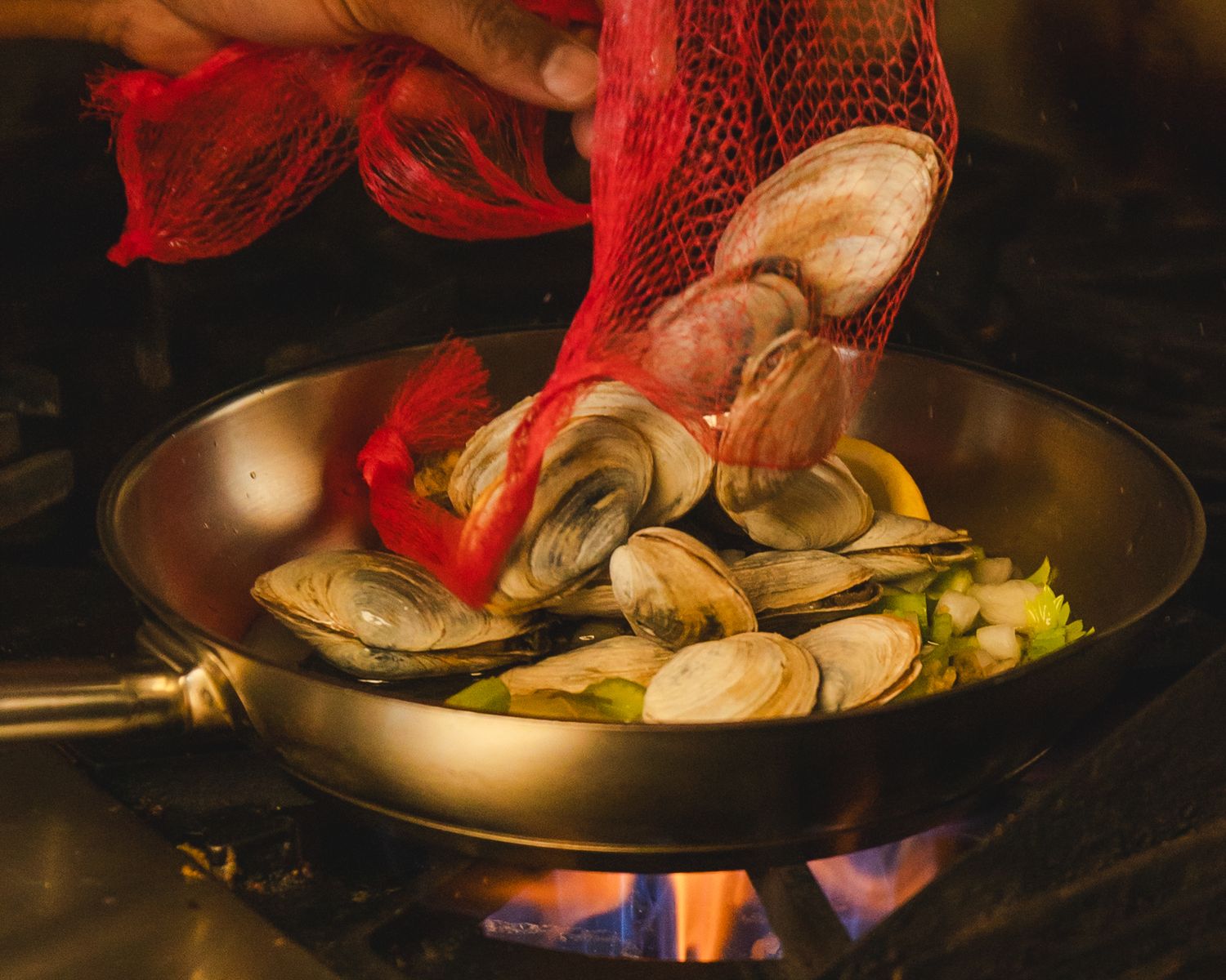 Clams being poured from a red mesh bag into a wok with vegetables over an open flame.