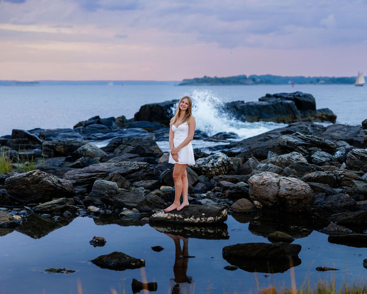 Young woman in white dress standing on coastal rocks at sunset with ocean waves crashing behind her.