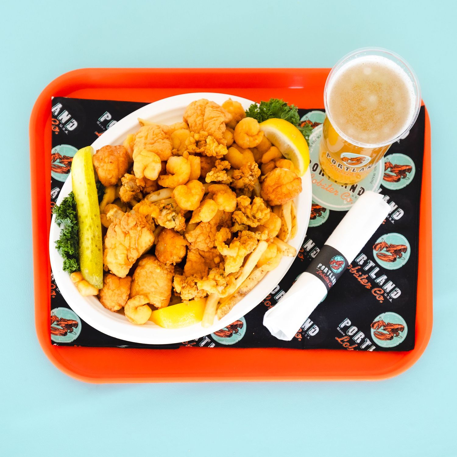 Fried seafood platter with lemon wedges, hush puppies, and a beer on an orange fast food tray.