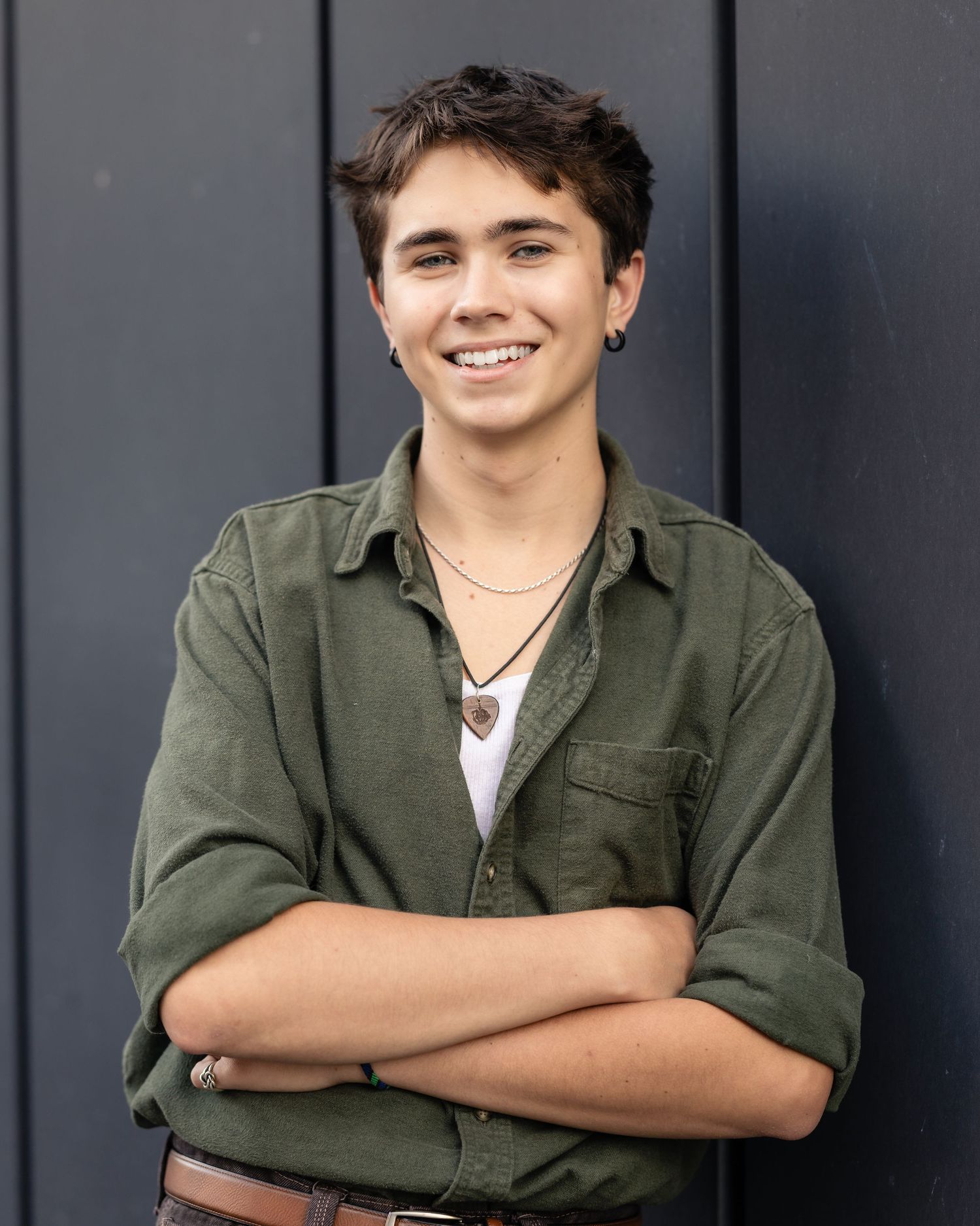 Smiling young person with crossed arms wearing a green shirt and necklace, posing against a dark wall.